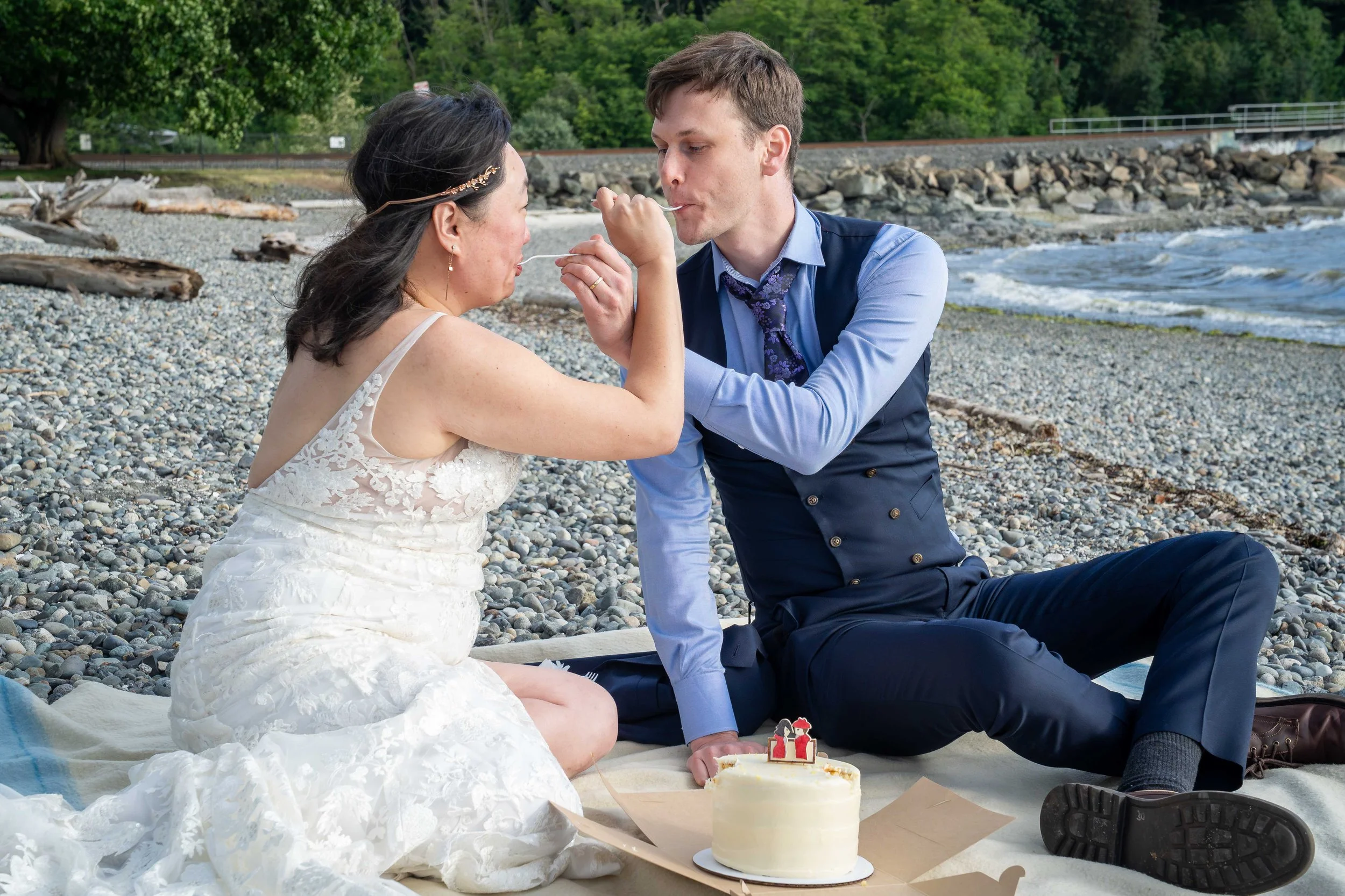 A photo of a bride and groom sitting on a picnic blanket on the beach at Marine Park in Bellingham with a Pure Bliss wedding cake between them. They are each feeding the other a piece of cake with their arms intertwined.