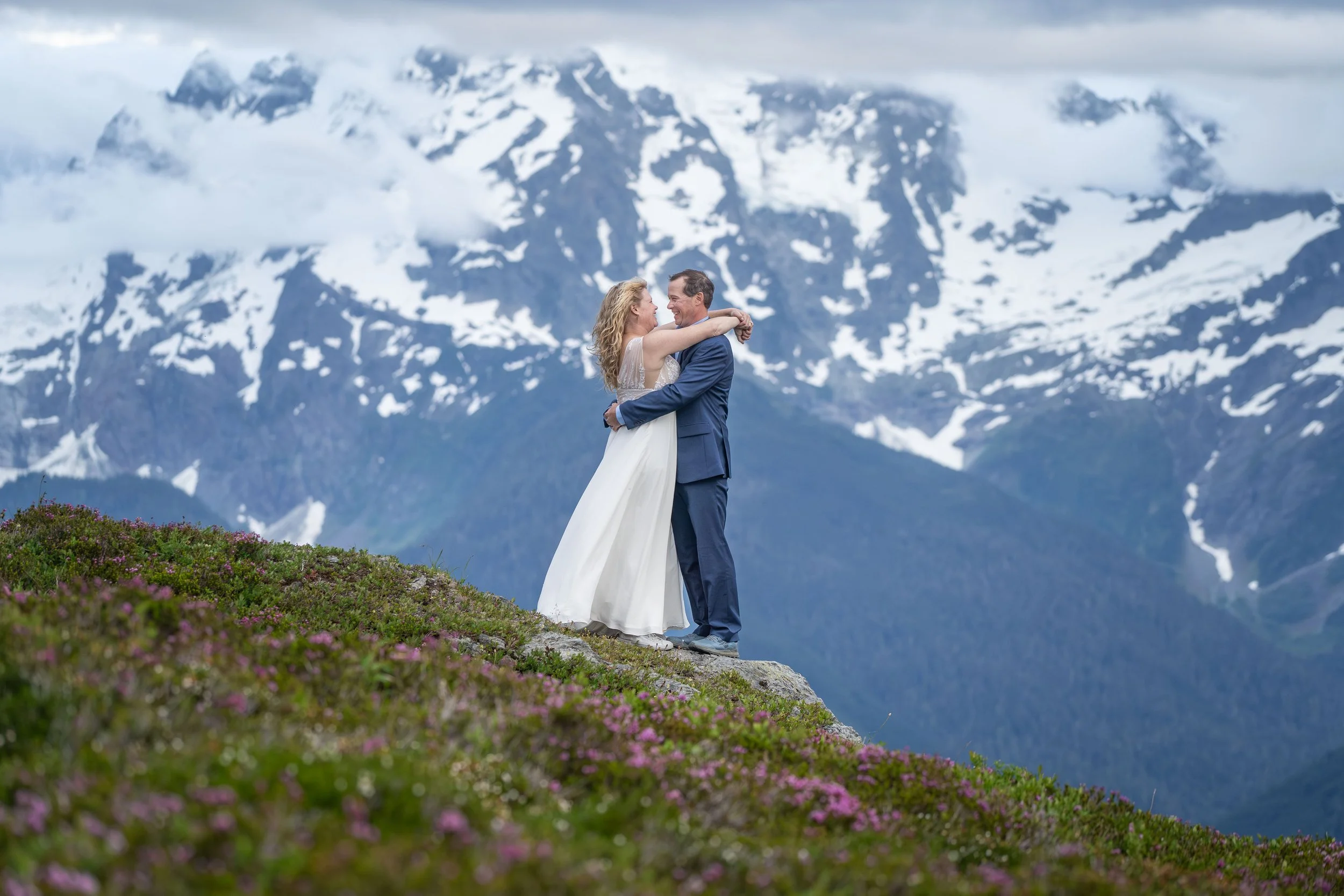 A bride and groom stand together at sunset in a subalpine meadow on Yellow Aster Butte after their North Cascades elopement, with Mt. Shuksan visible behind. The photo uses telephoto compression to make the snowy face of Shuksan appear very large..