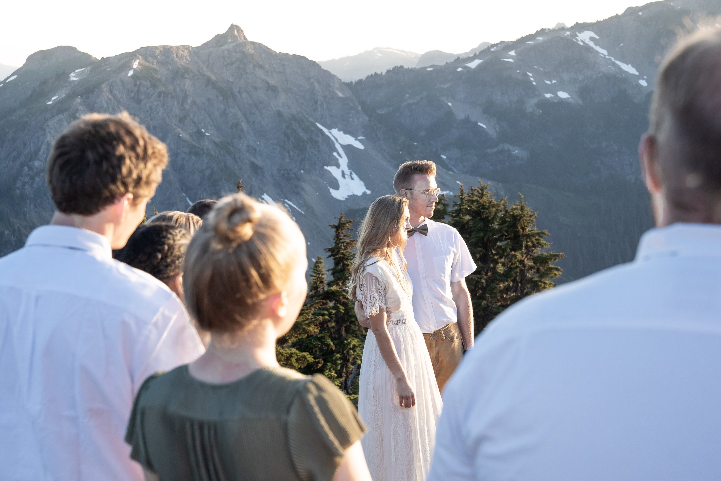 A photo of a couple in wedding clothes getting married in the North Cascades with large mountains behind them. The photo is taken from behind the bridesmaids and groomsmen so that the couple is visible between them.