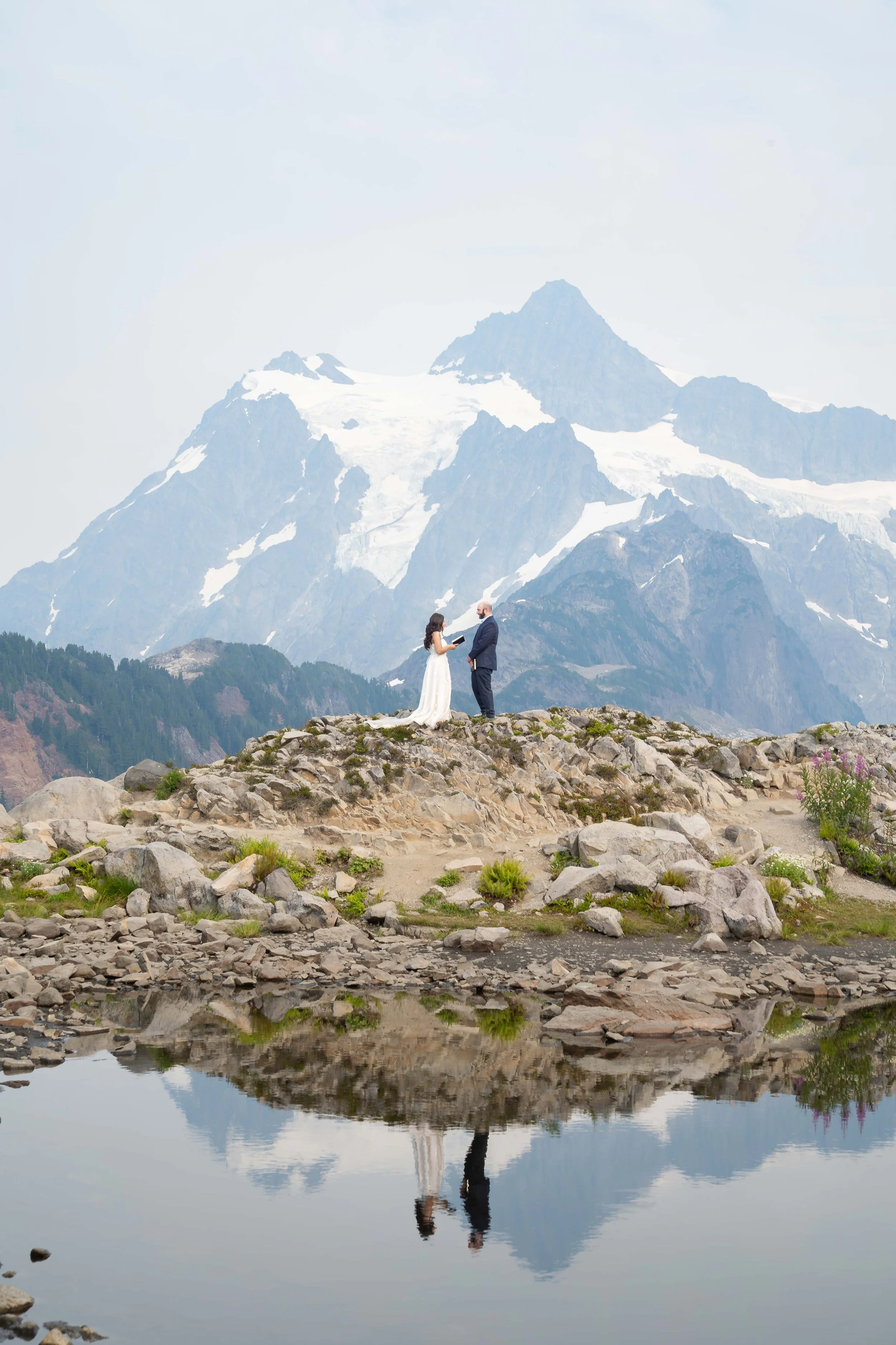 A bride and groom reading their vows during an elopement ceremony at Artist Point, directly in front of Mt. Shuksan. They are standing above an alpine lake that mirrors their reflection. It's smoky, creating a haze over the mountain.