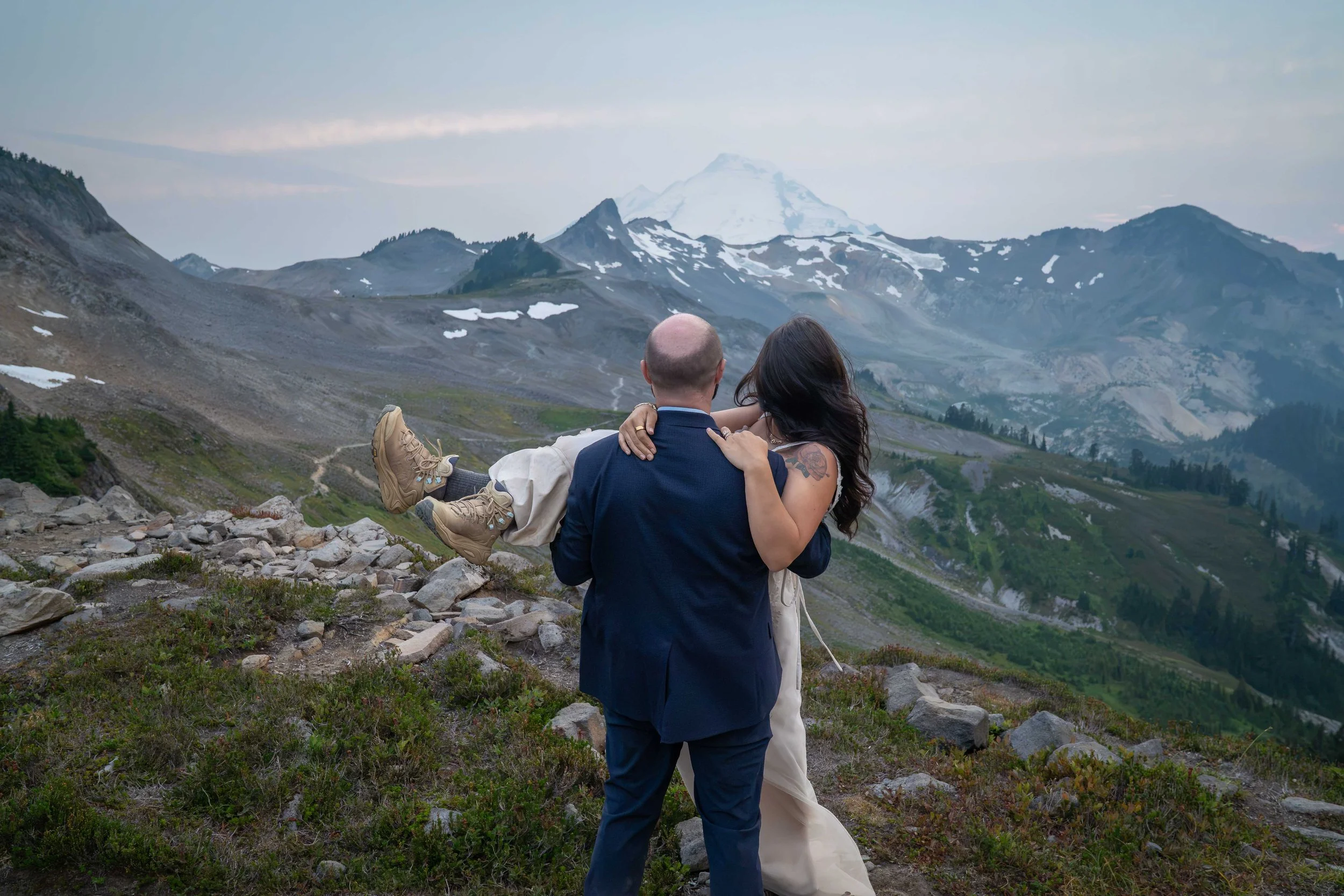 A groom holds his bride in his arms at sunset as they look towards Mt. Baker at the end of their Ptarmigan Ridge elopement. The bride's hiking boots are visible, and the photo shows the scale of the landscape including Coleman Pinnacle and Mt. Baker.