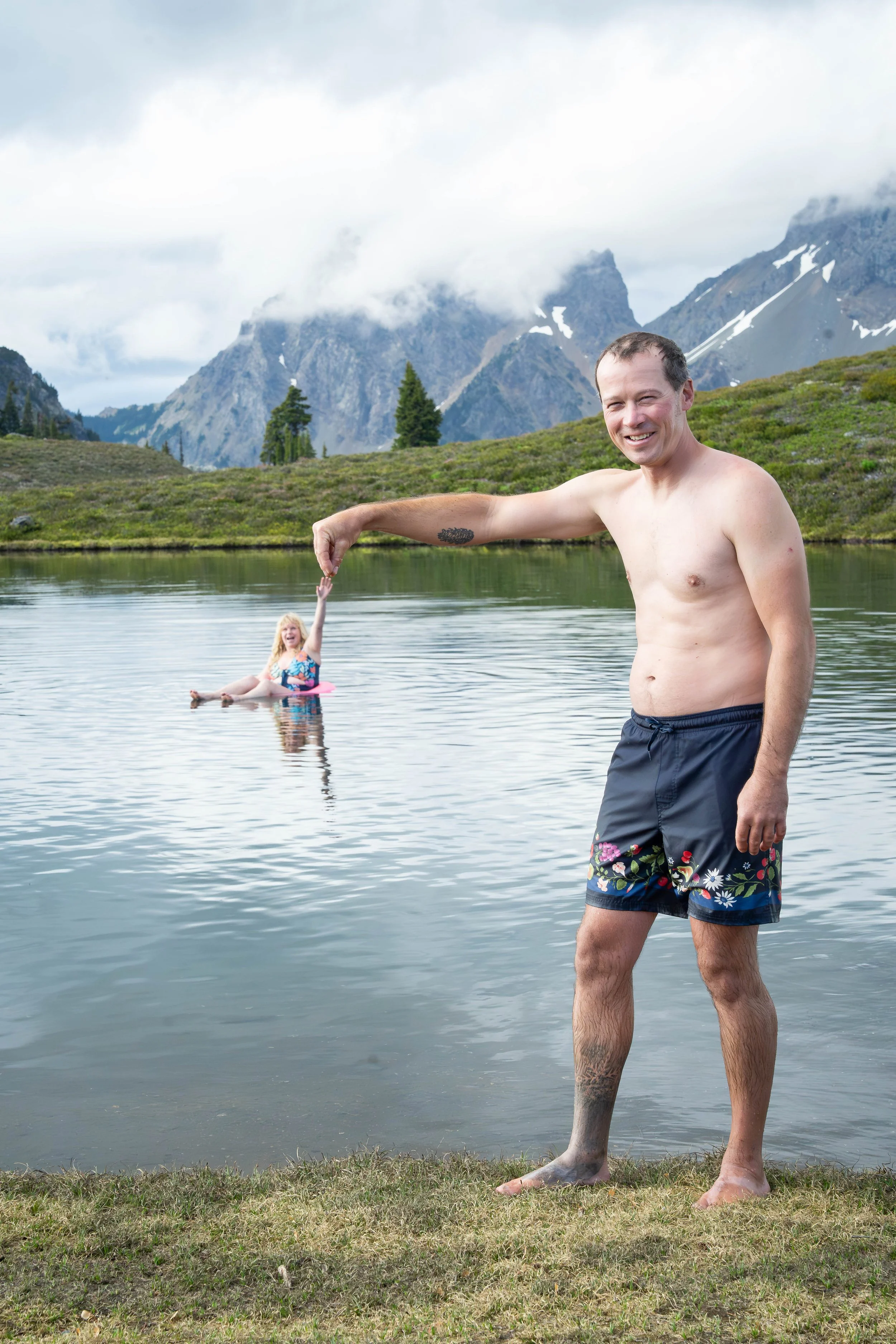 A telephoto compression photo of a groom picking up his bride by the hand. The groom is wearing a bathing suit and the bride is floating on a pink innertube in an alpine lake in the North Cascades. American Border Peak is visible in the background.