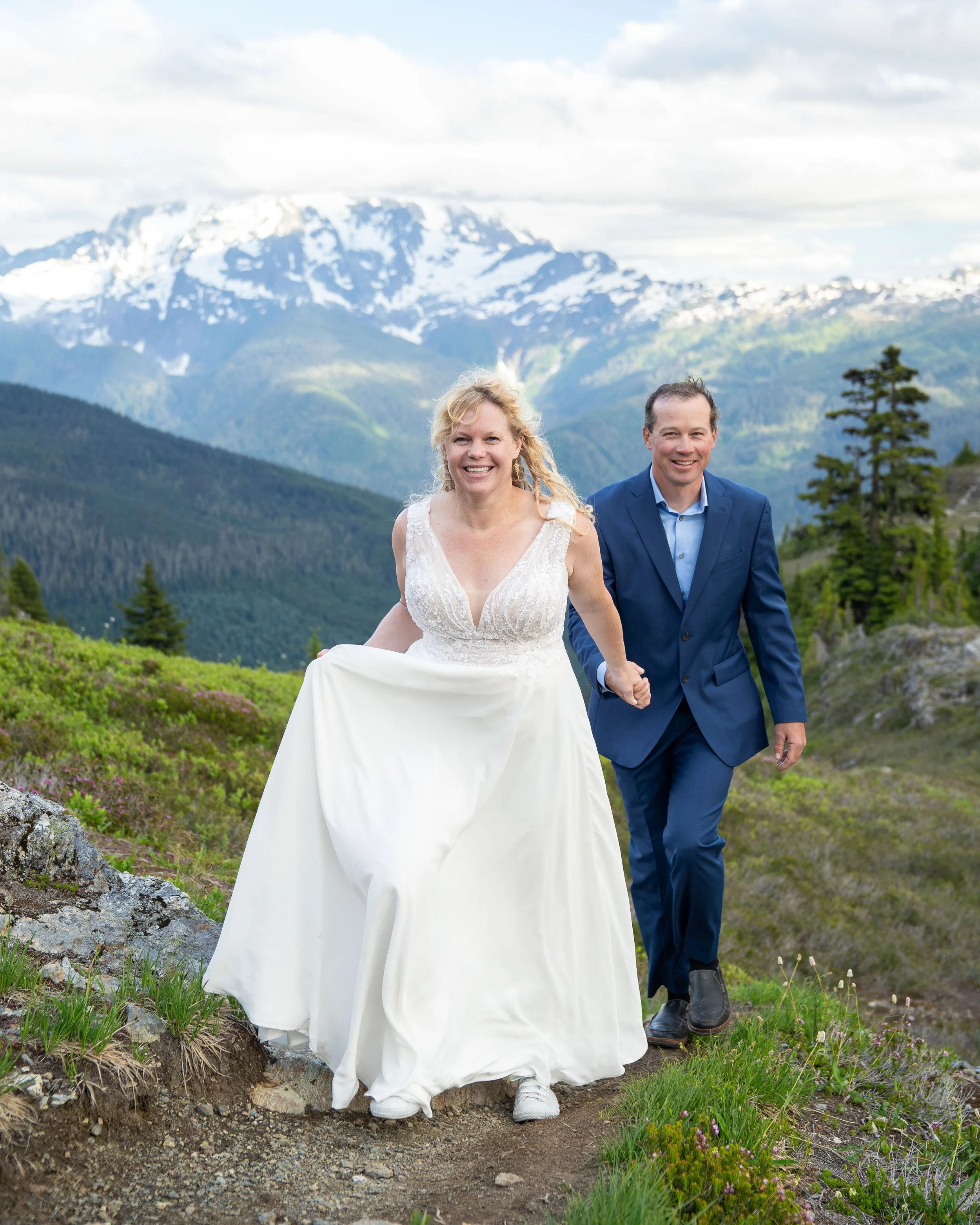 A bride and groom in wedding clothes walking towards the camera in a subalpine meadow with blooming wildflowers and Mt. Baker behind them. The couple is eloping at Yellow Aster Butte in the North Cascades.