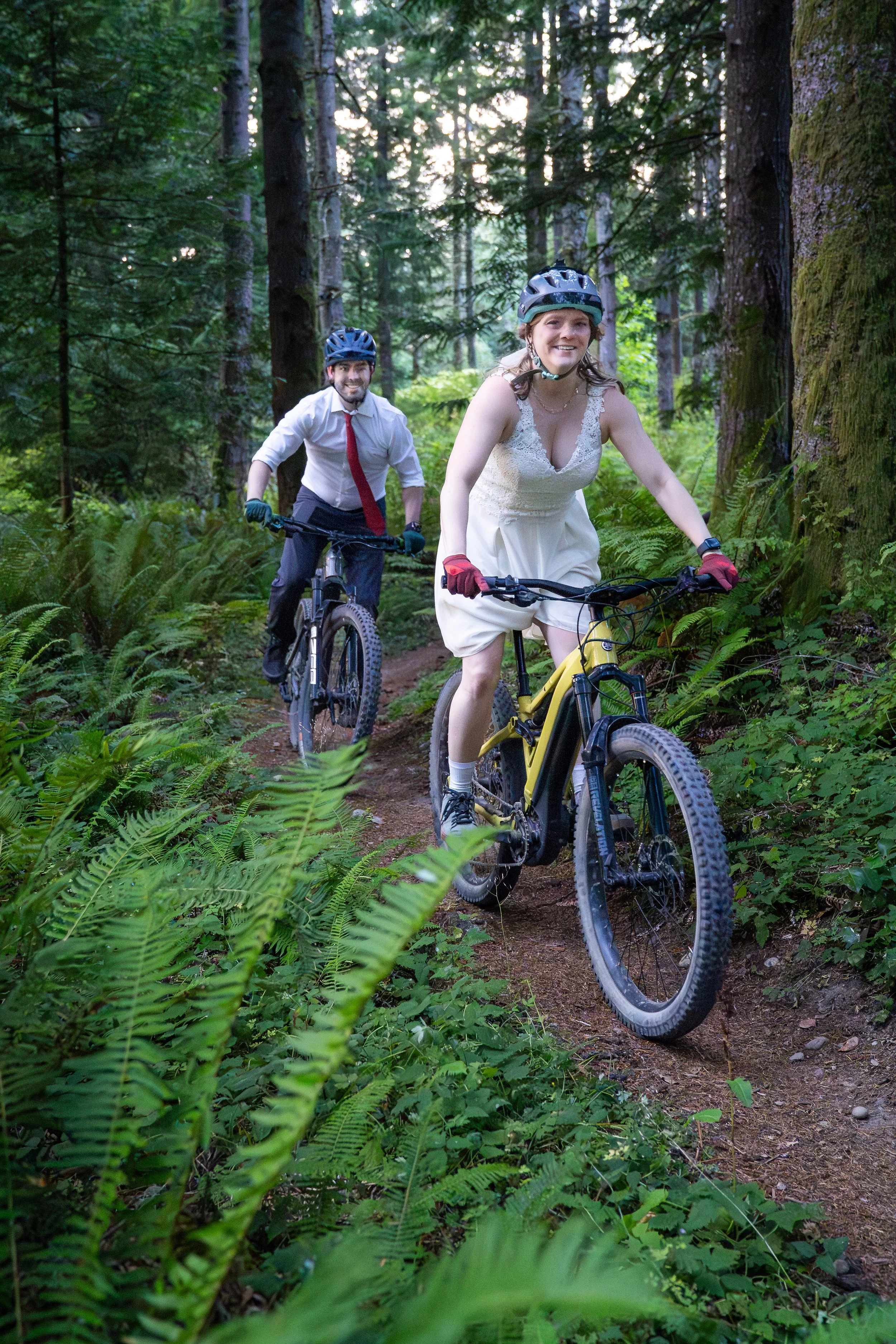 A bride and groom riding mountain bikes in full wedding clothes during a mountain biking elopement photoshoot on Galbraith Mountain, in Bellingham, Washington. They are on singletrack surrounded by ferns and mossy trees.