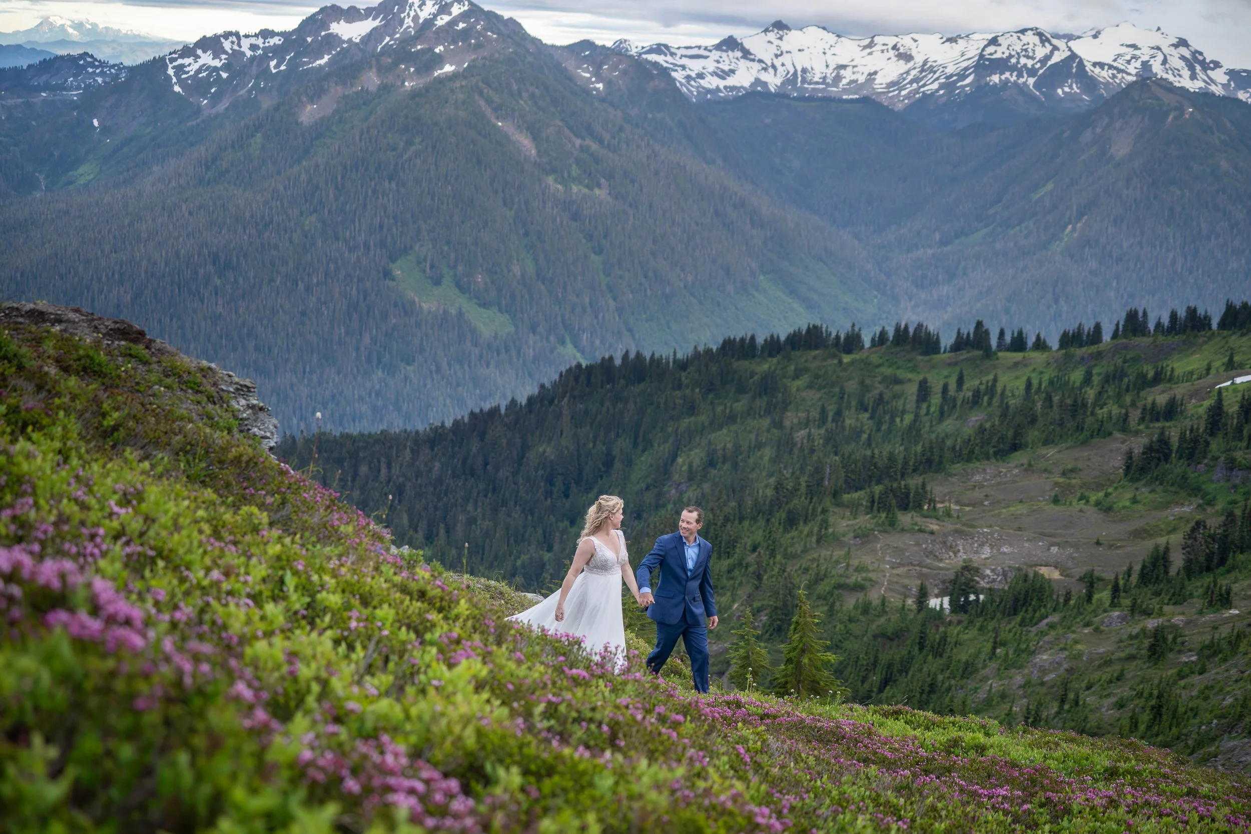 A bride and groom walking together through an alpine meadow of blooming wildflowers and purple heather during an elopement at Yellow Aster Butte in the North Cascades.