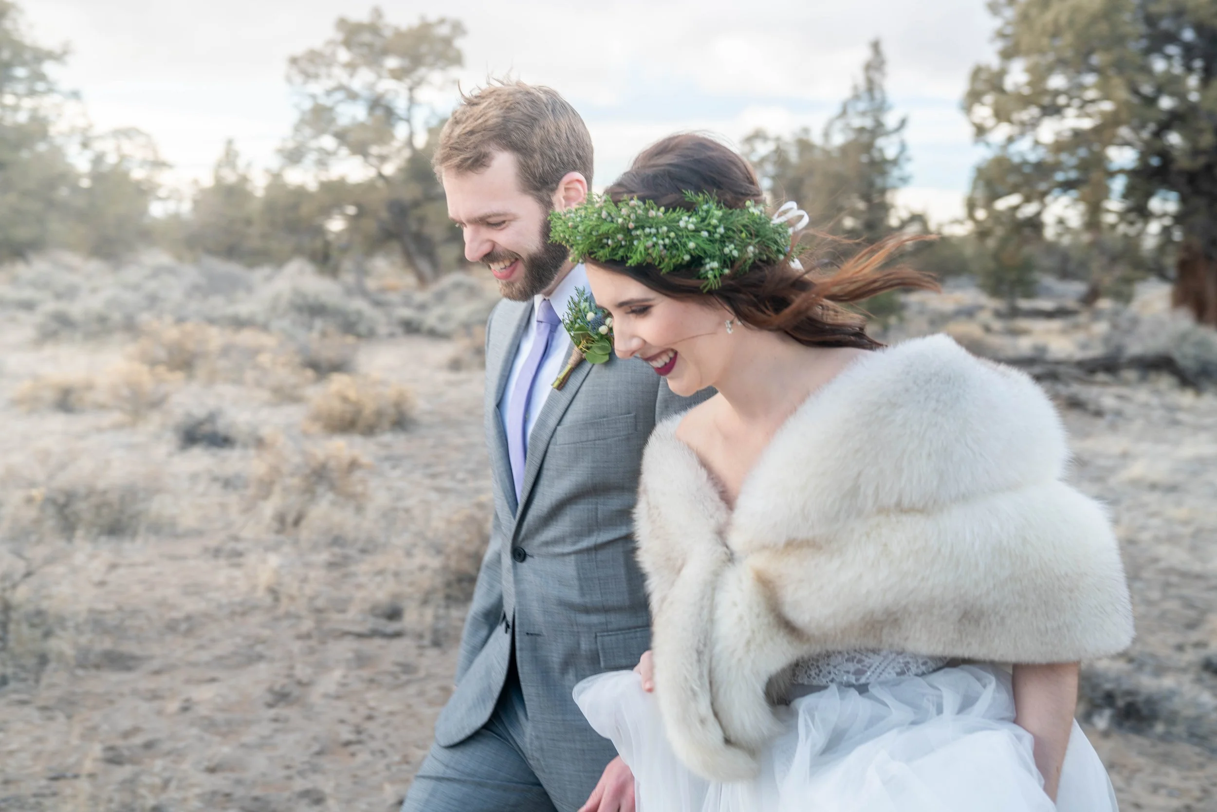 A couple in wedding clothes walking together through a sagebrush desert. The bride is wearing a wedding gown and fur coat, and the groom is wearing a suit.