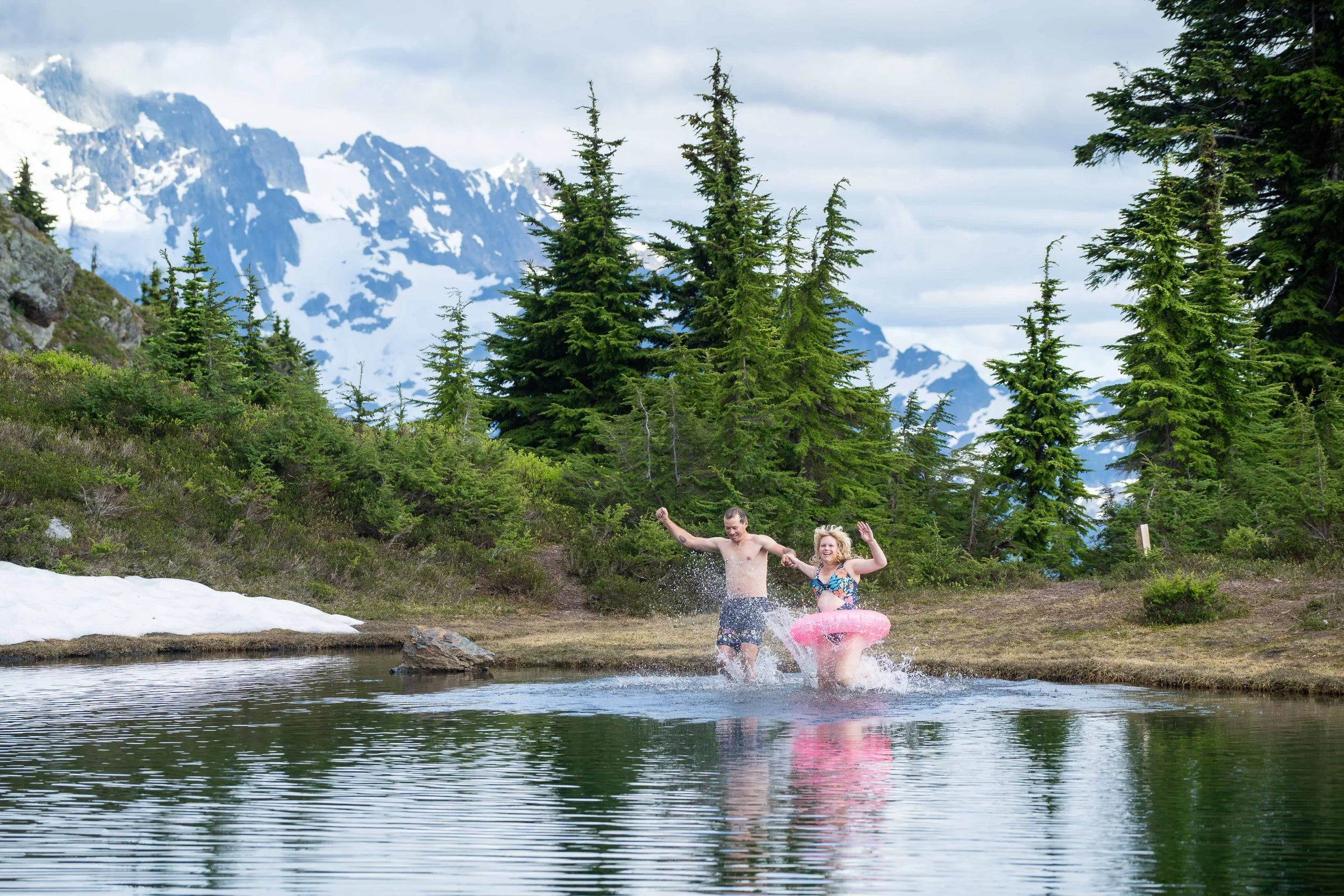 A bride and groom running and splashing into an alpine lake around Yellow Aster Butte during their North Cascades elopement. There's snow on the edge of the lake and Mt. Shuksan is visible in the background.
