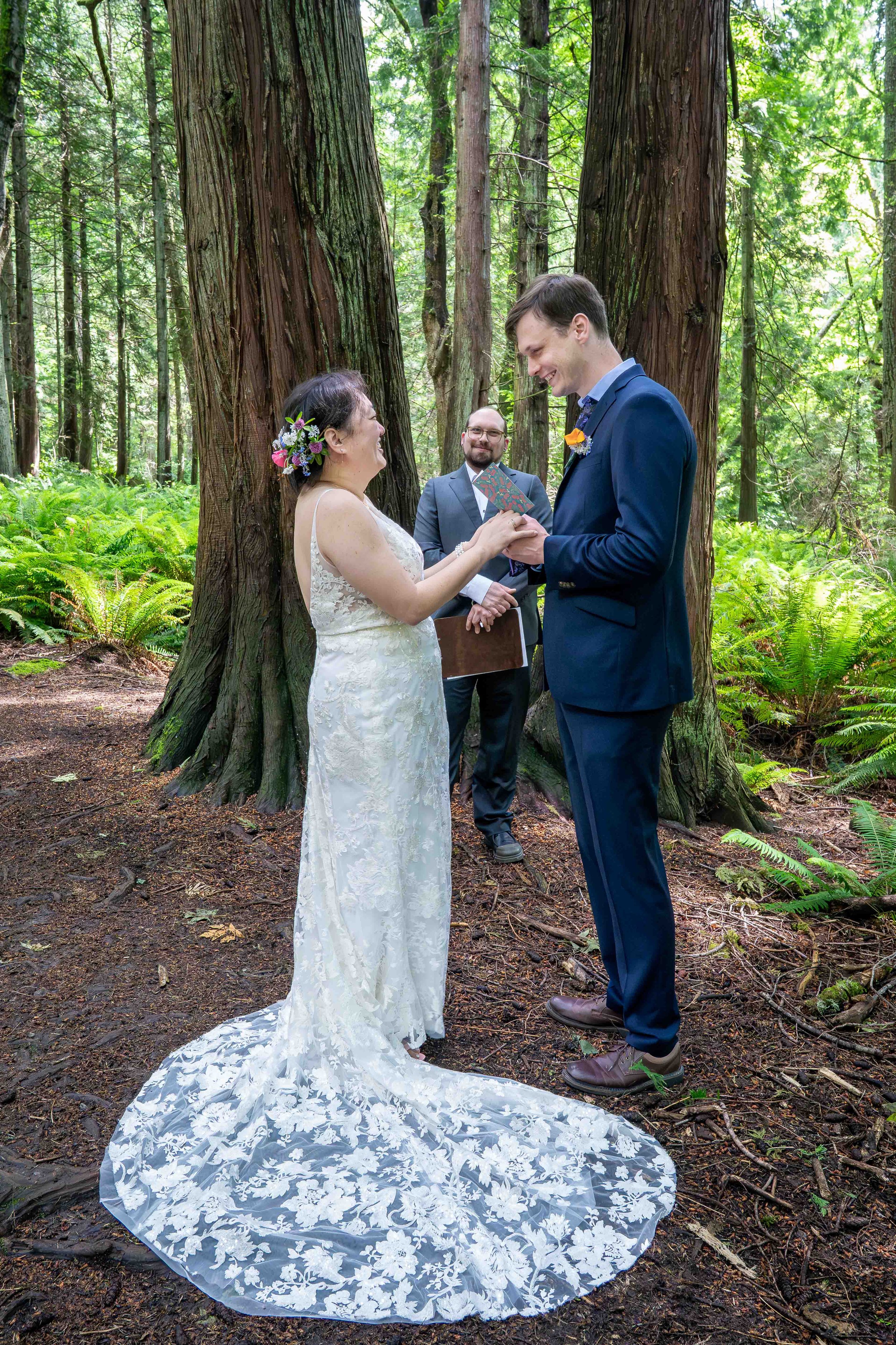 A bride and groom smile at each other as the groom reads his vows and their officiant looks on during an elopement wedding ceremony. They are surrounded by large cedars and ferns in the lush forest of the Fragrance Lake Trail in Larrabee State Park.