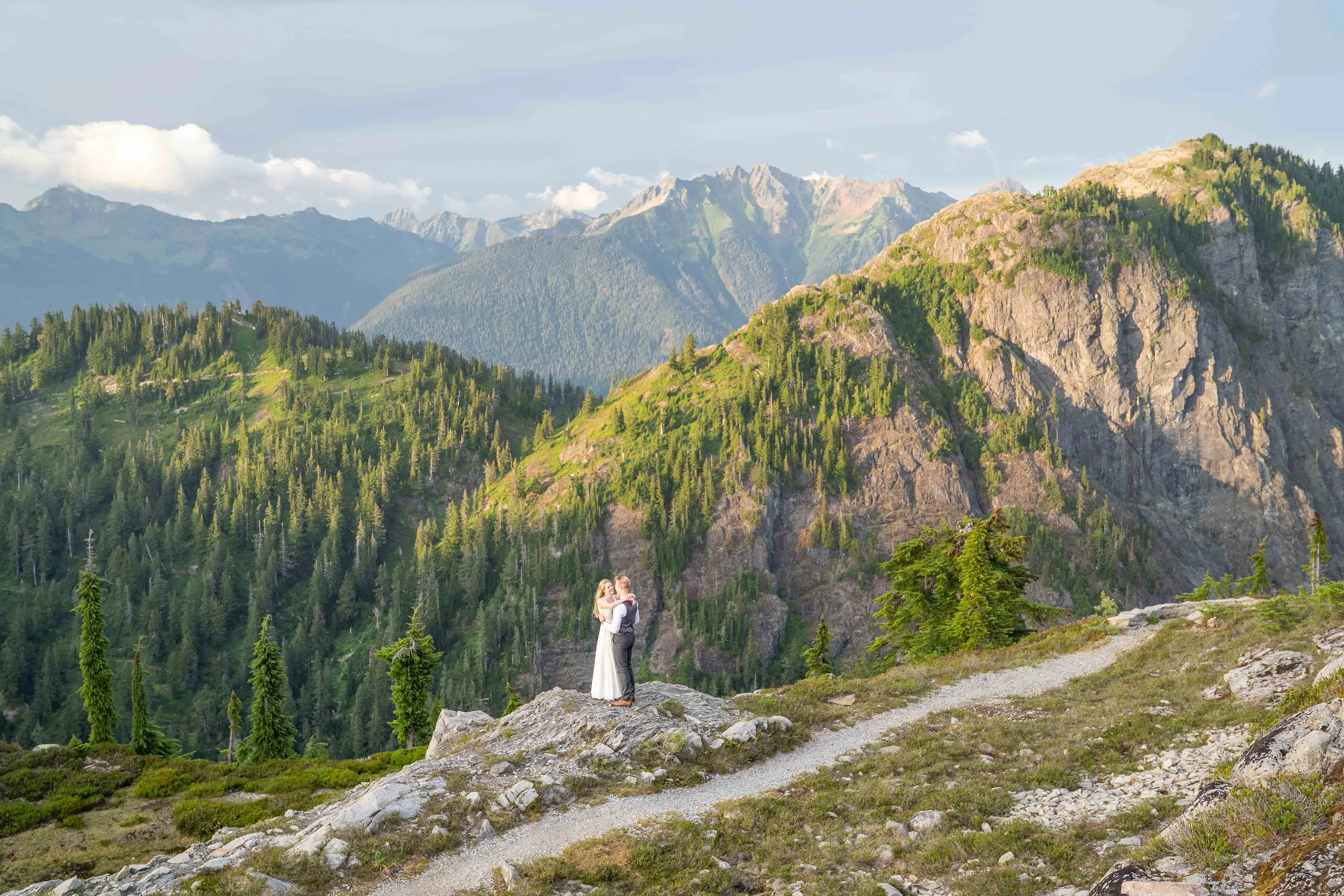 Photo of a bride and groom standing together during an Artist Point, Washington elopement at sunset. They're standing on a rock with the trail visible in front of them and the rocky peaks of the North Cascades behind them.