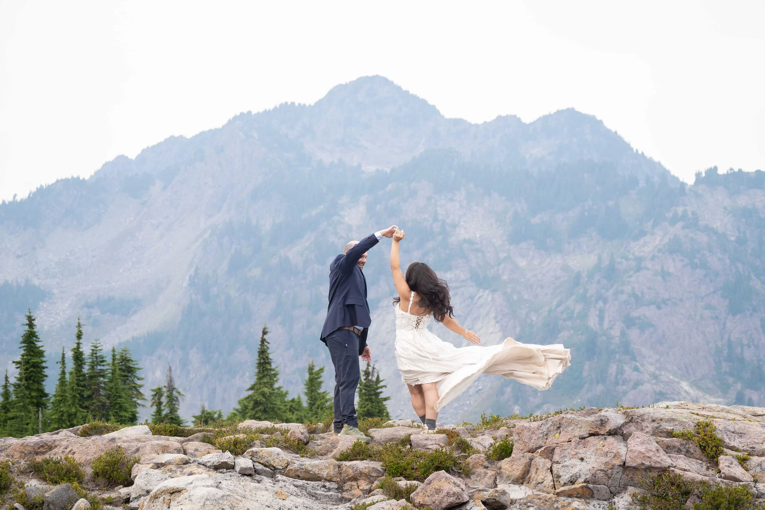 A groom spins his bride, whose dress is flowing through the air behind her, during a Mt. Baker elopement at Artist Point. They are standing in a rocky alpine meadow and Mt. Herman and the North Cascades are visible behind them.