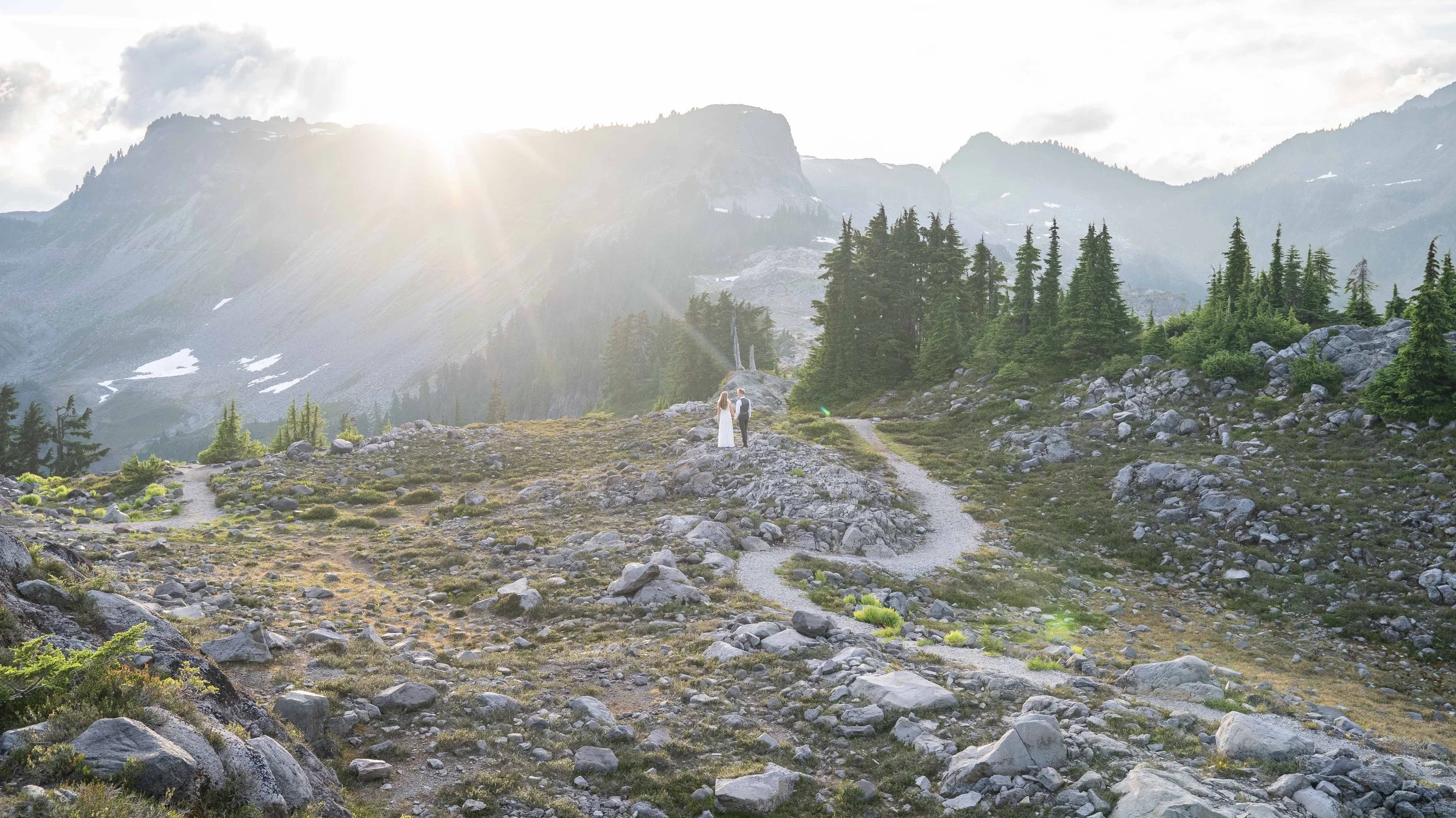 A photo of a bride and groom in wedding clothes standing together on rocks next to a trail at Artist Point during their elopement. They're watching sunset over Table Mountain and they're surrounded by an alpine meadow.