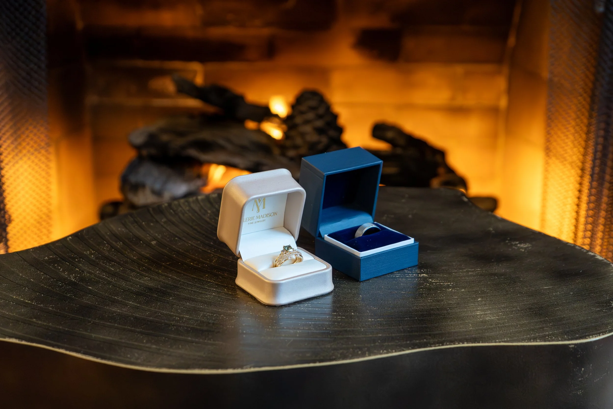 Photo of a pair of wedding rings in ring boxes on a dark wooden table in front of a lit fireplace.