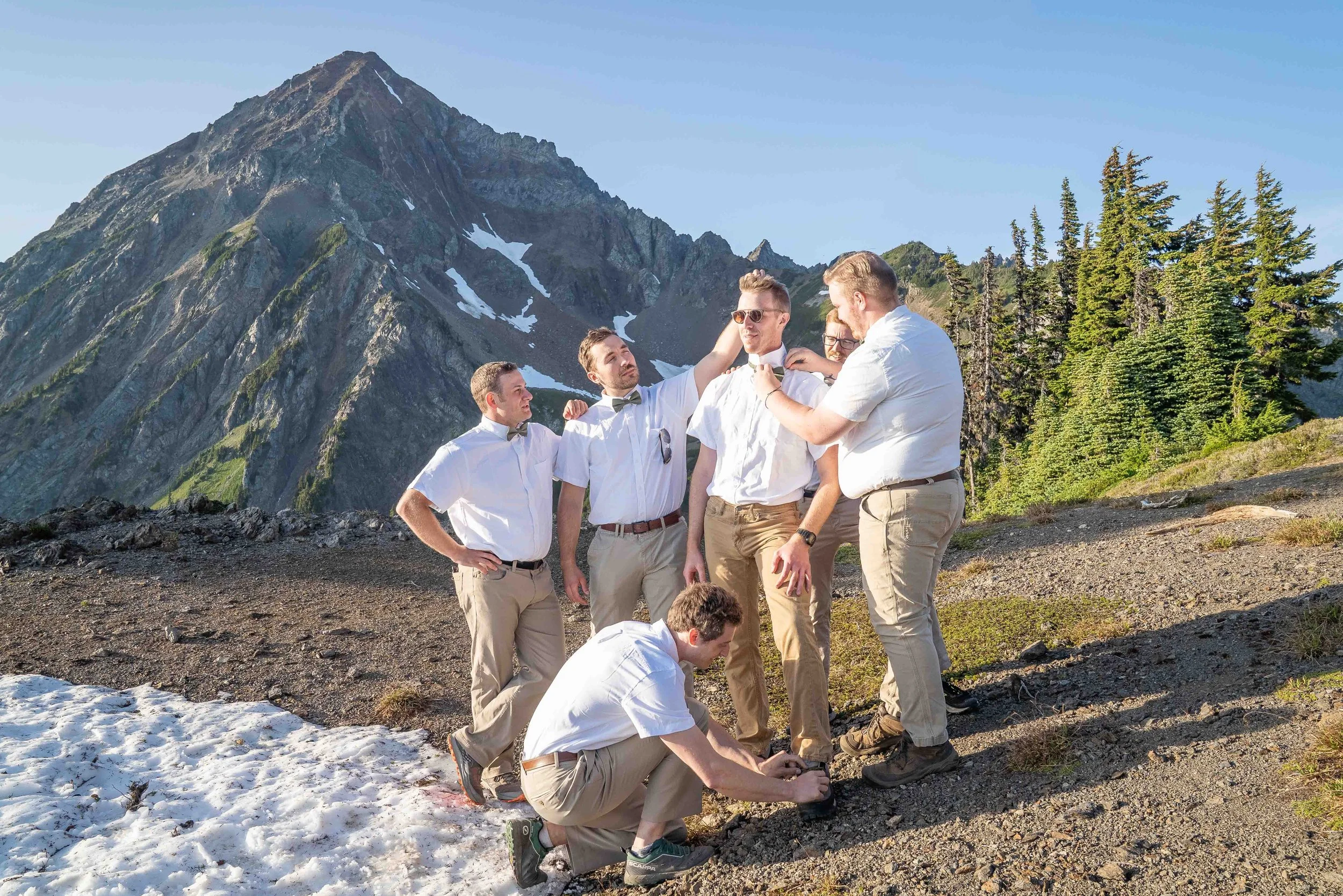 A group photo of a groom in his wedding clothes being attended to by his groomsmen in formal wear during a vow exchange at High Pass in the North Cascades, Washington. Mount Larrabee is directly behind them.