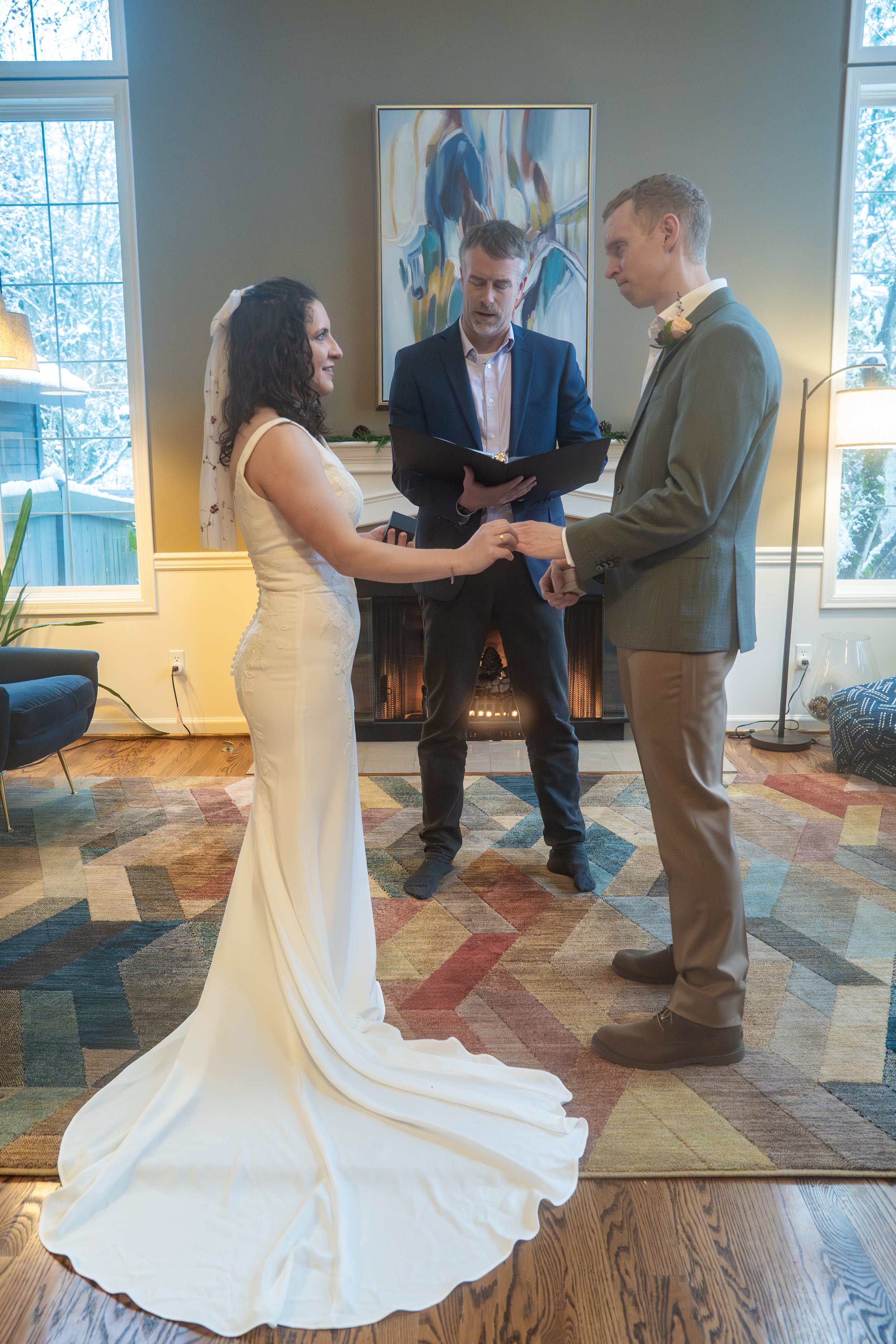 Photo of a couple in the middle of a wedding ceremony, as the woman puts a wedding ring on the man's hand. They're in a living room in front of a fireplace, with the officiant between them.