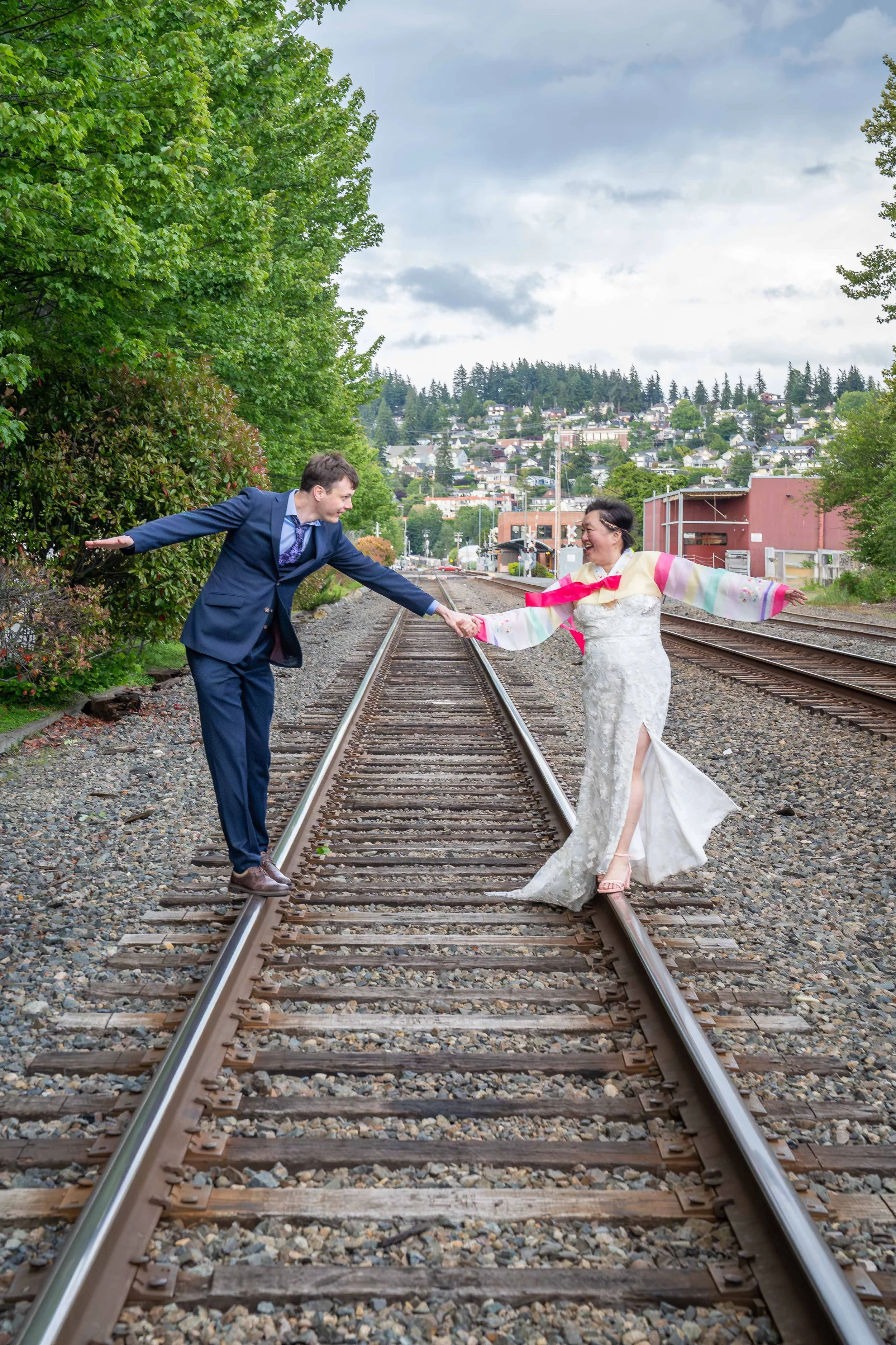 A photo of a bride and groom after their elopement standing on railroad tracks and holding hands, pulling on each other for balance as they lean backwards. The tracks run into the distance behind them with the city of Bellingham in the background.