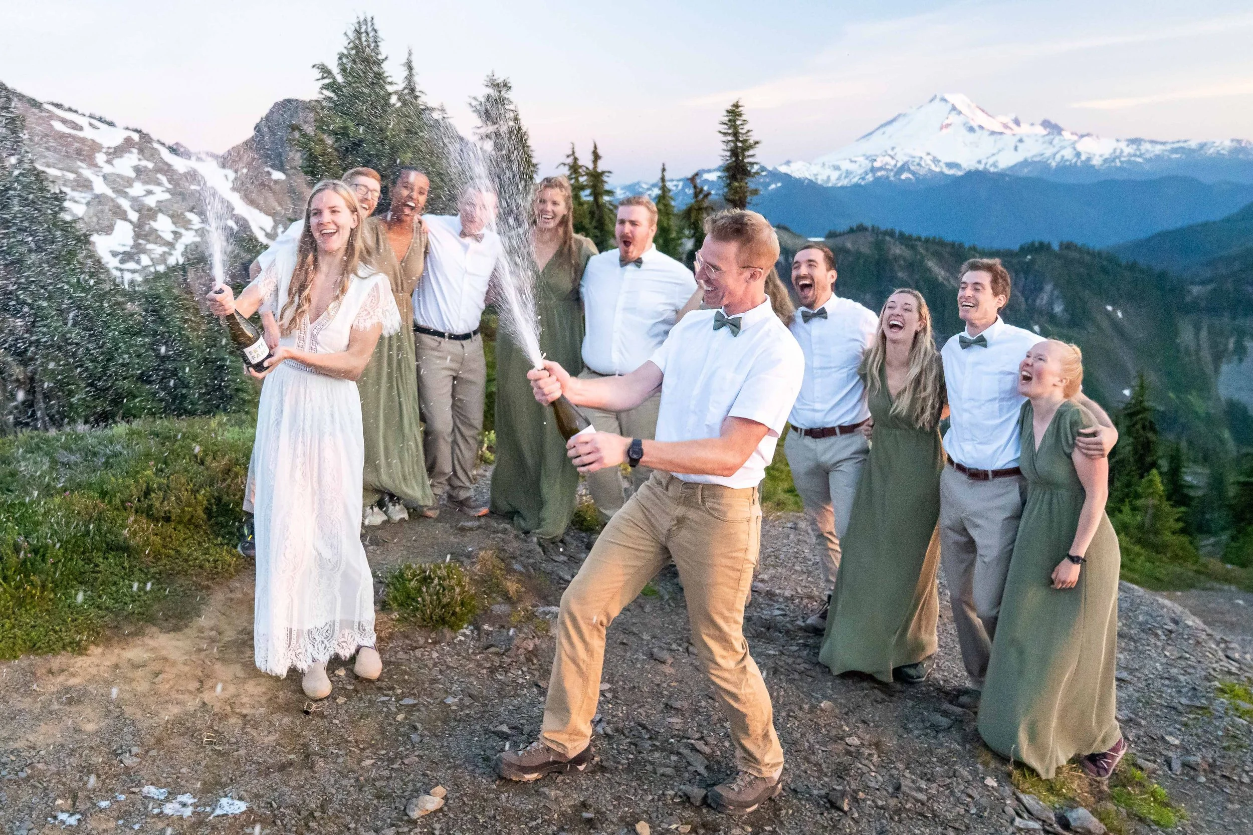 A photo of a just-married bride and groom popping champagne bottles after a backcountry elopement in the North Cascades in Washington. The couple is surrounded by their bridesmaids and groomsmen and Mt. Baker is visible in the background.