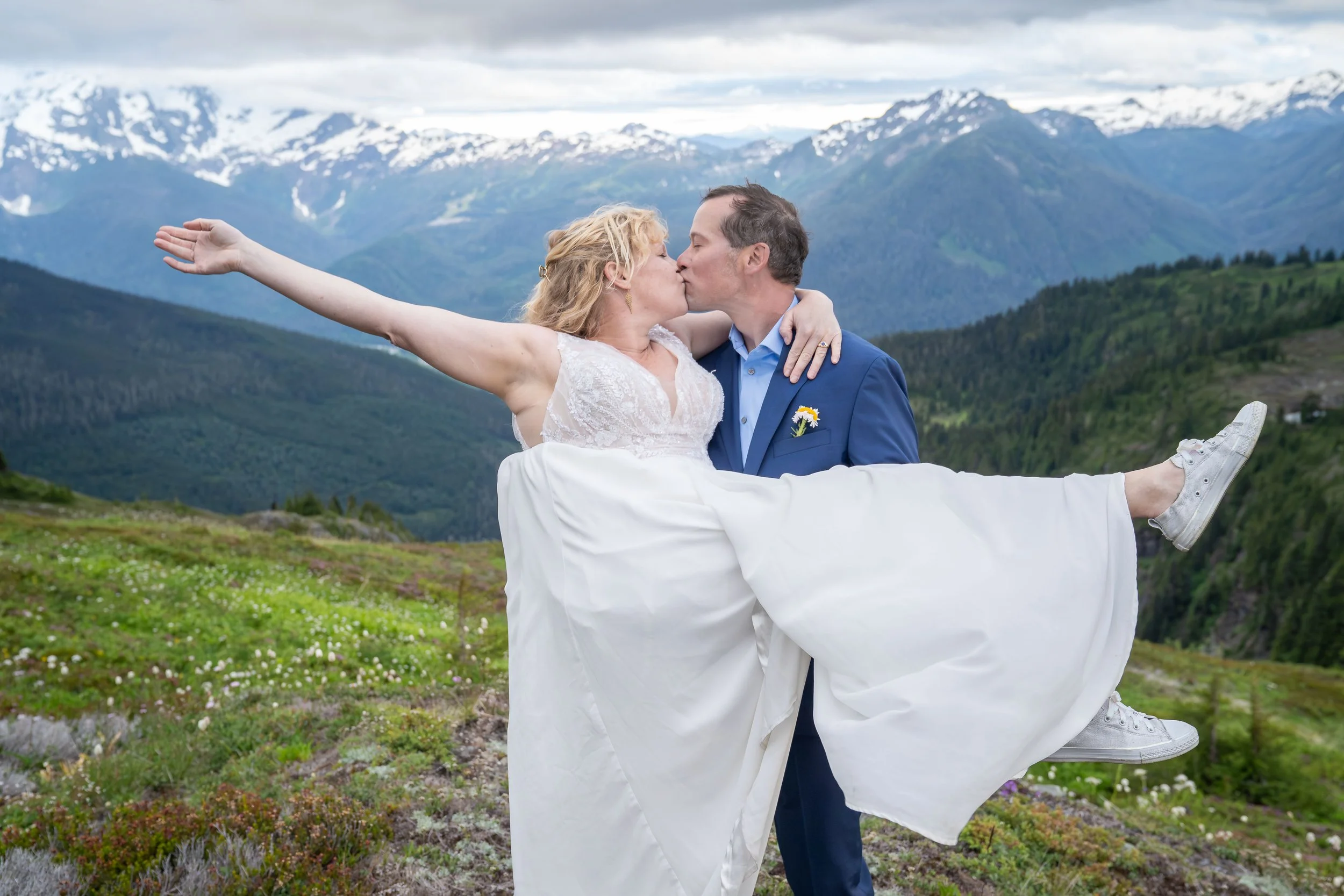 A groom holding his bride in the air during a North Cascades elopement at Yellow Aster Butte, with Mt. Shuksan and Mt. Baker visible in the background. The couple is standing in a subalpine meadows with blooming white and purple wildflowers.