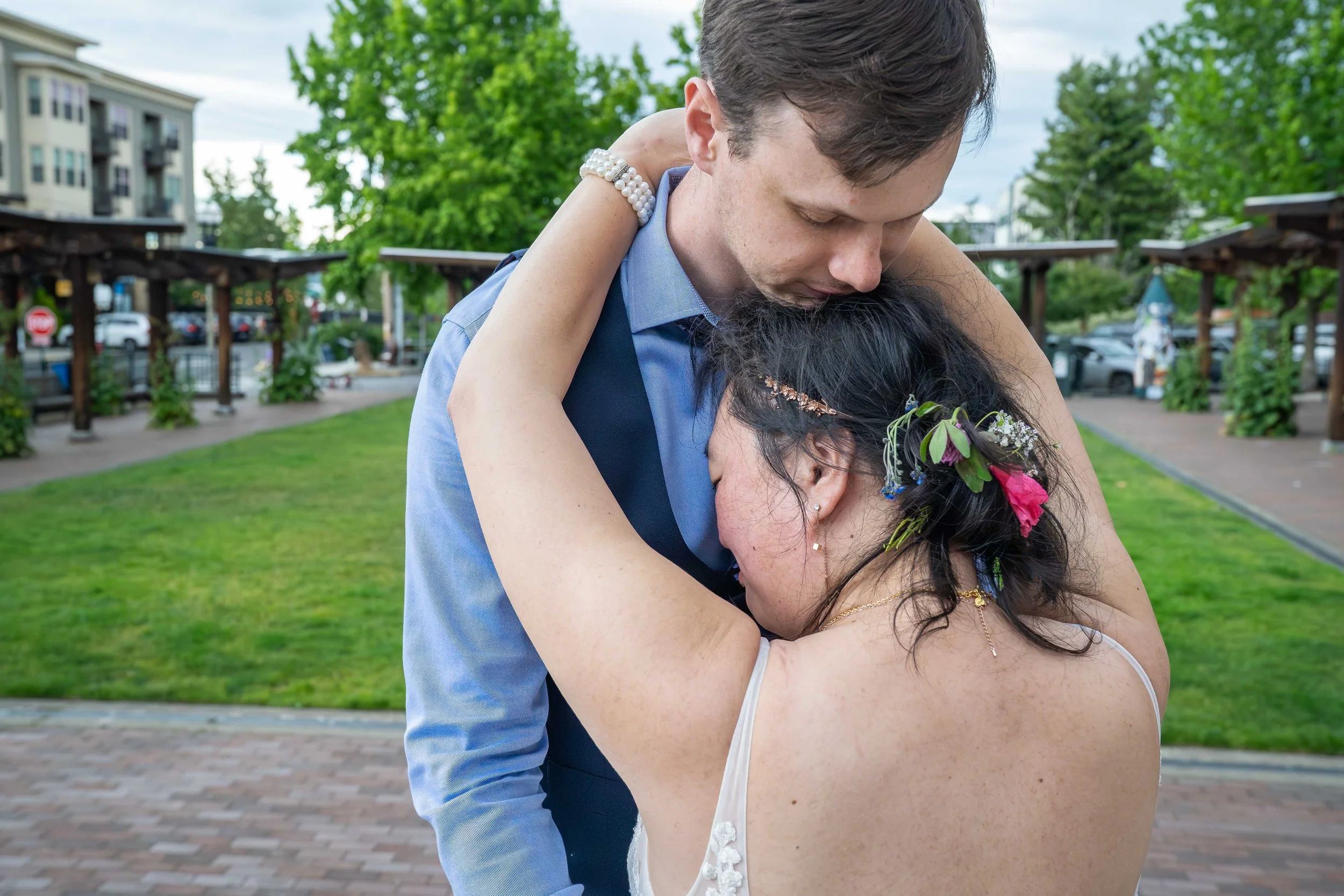 A bride puts her arms around her groom's shoulders and presses her face into his chest after their Bellingham elopement. They are in the Village Green in downtown Fairhaven and the brick-lined plaza is visible behind them.