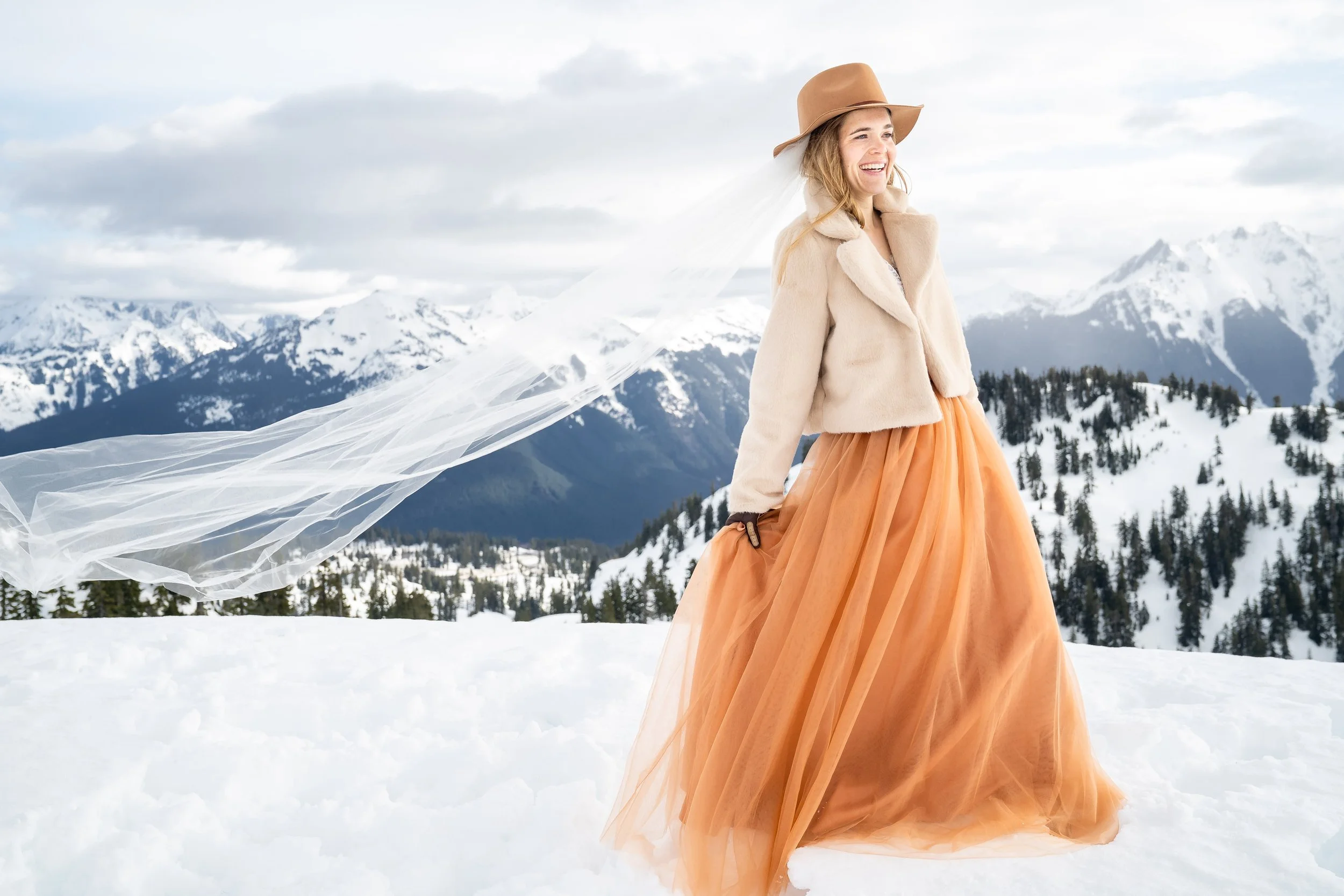 A photo of a bride wearing an orange dress, white fur coat, and hat standing in the snow with mountains behind her and her veil blowing in the wind.