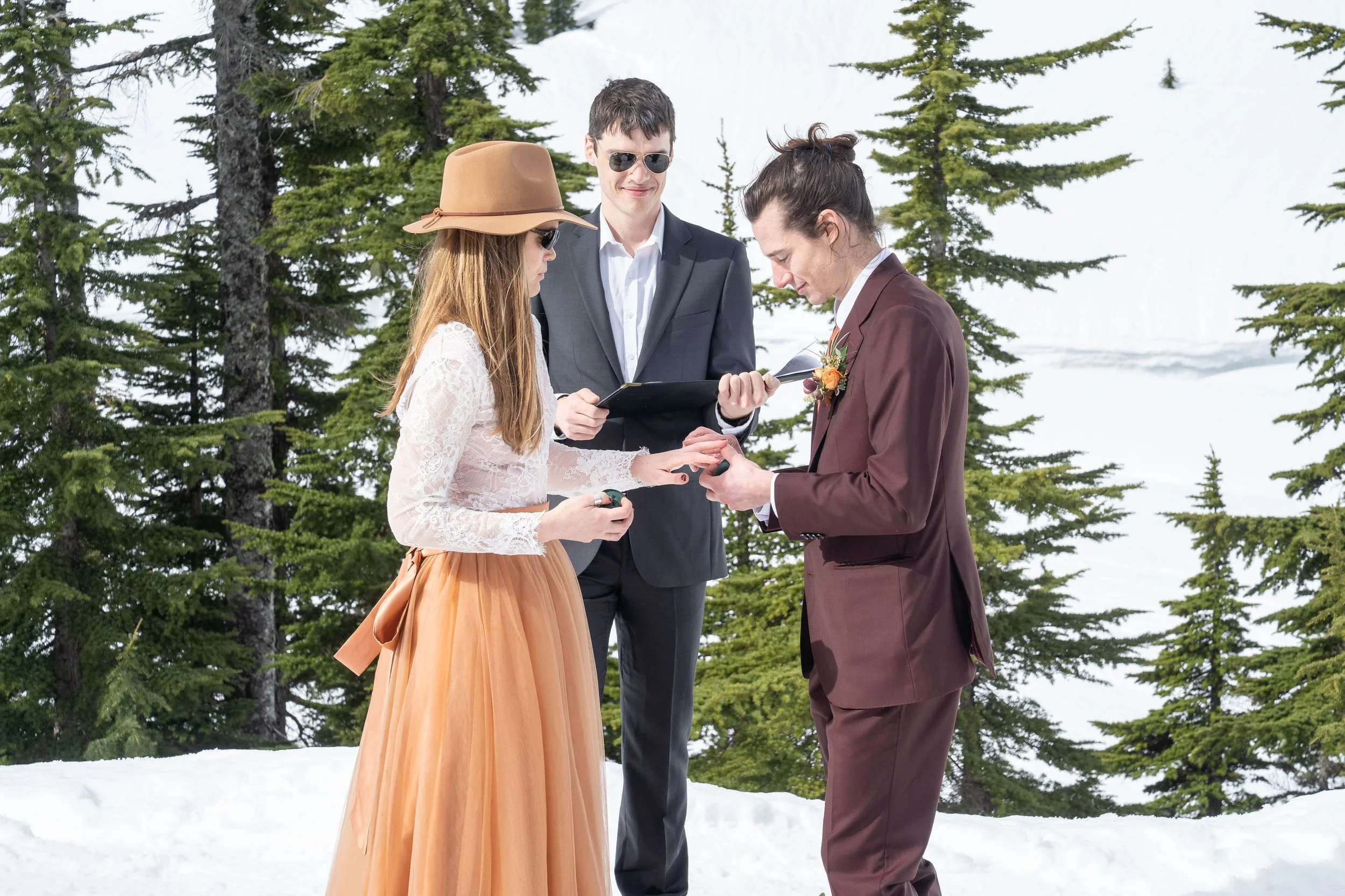 A photo of a wedding ceremony in the snow at Heather Meadows. The groom is placing a ring on the bride's finger while the officiant looks on.