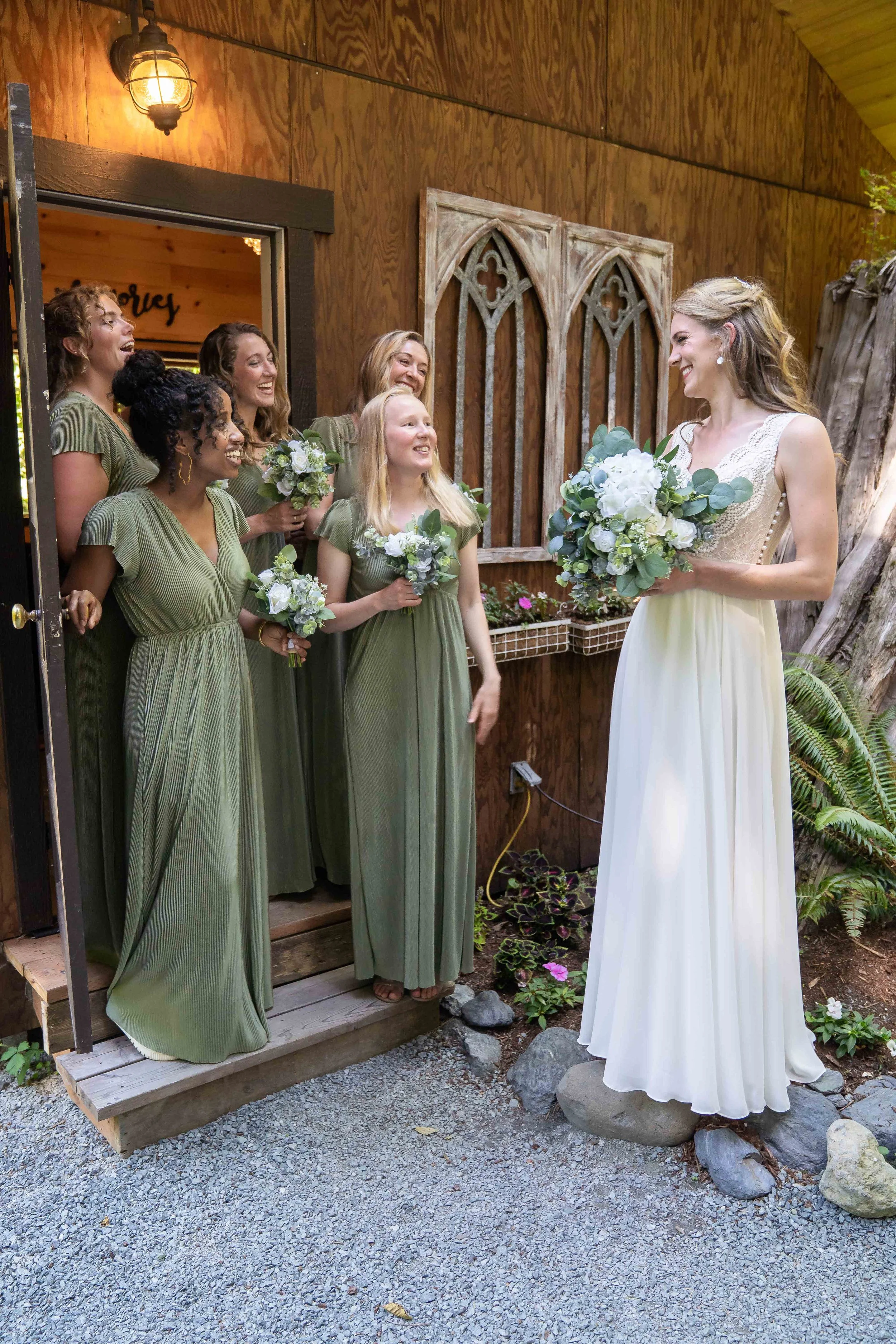 Photo of a bride and her bridesmaids at a wedding at Creekside Celebrations in Skagit County, Washington. The bride is wearing her dress and holding a bouquet, looking at her bridesmaids who are standing together.