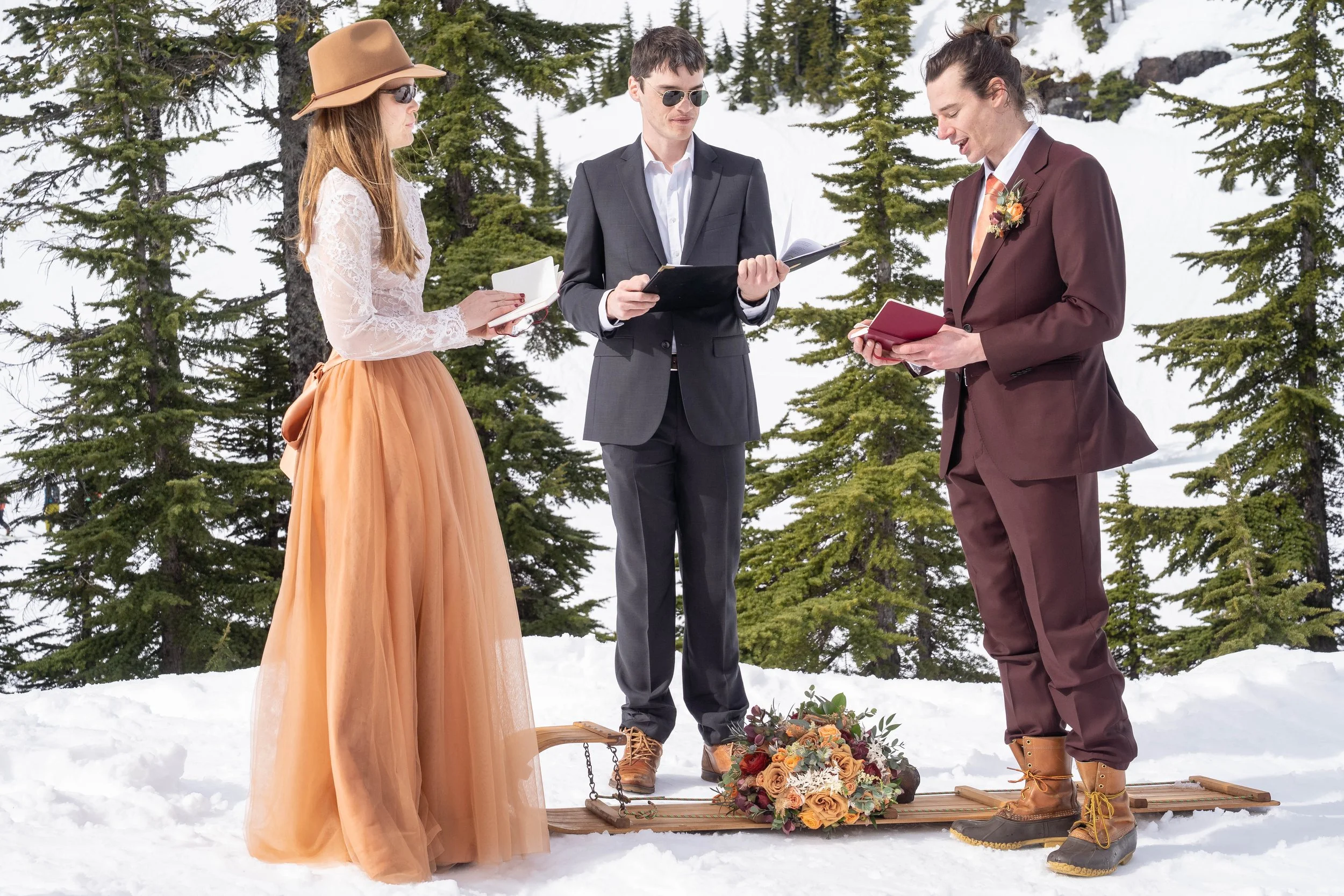 A photo of a wedding ceremony in the snow. The groom is reading his vows from a small notebook, while his bride and the officiant watch. There's a sled on the ground with a bouquet of orange flowers.