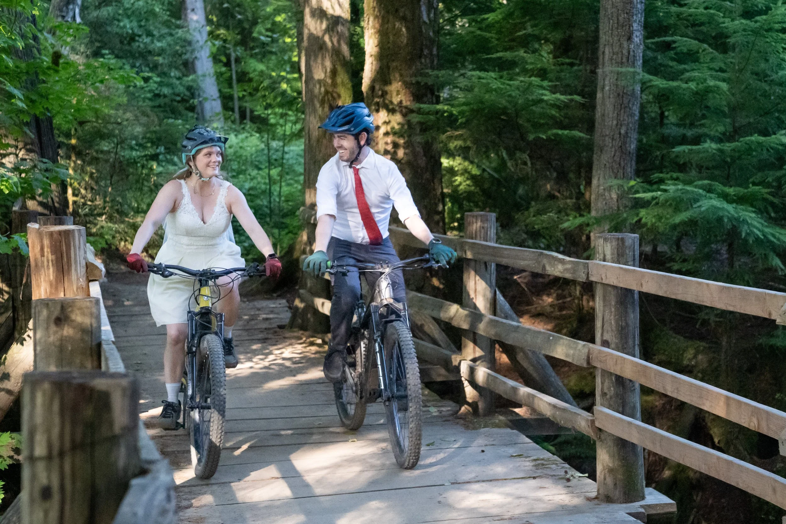 A bride and groom riding mountain bikes while wearing wedding clothes during a Galbraith Mountain elopement. They are riding over a wooden bridge on the trail Gate and Switch.