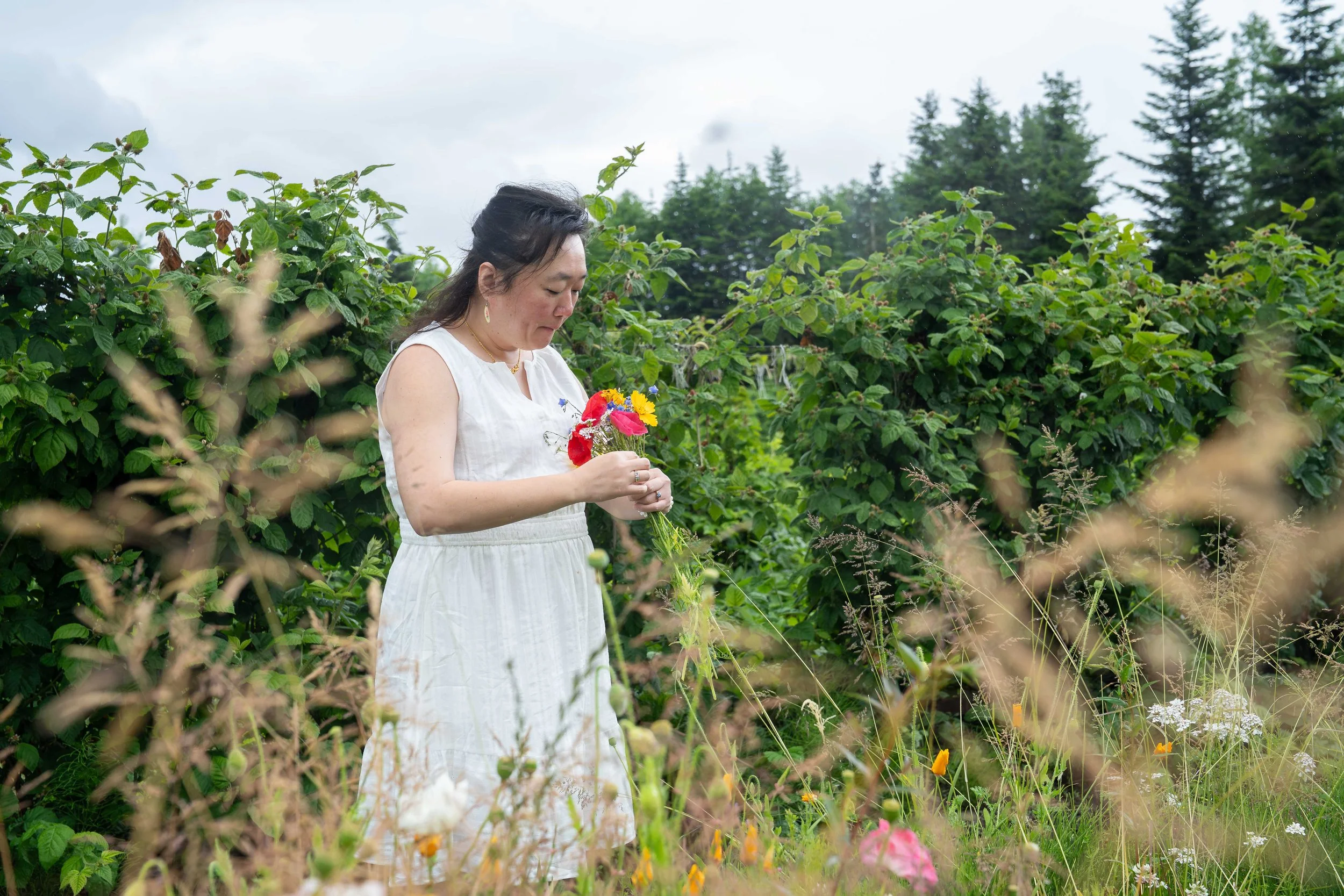 A bride in a white sundress standing in a field of flowers and holding a bouquet she has picked during a Bellingham elopement photography session.