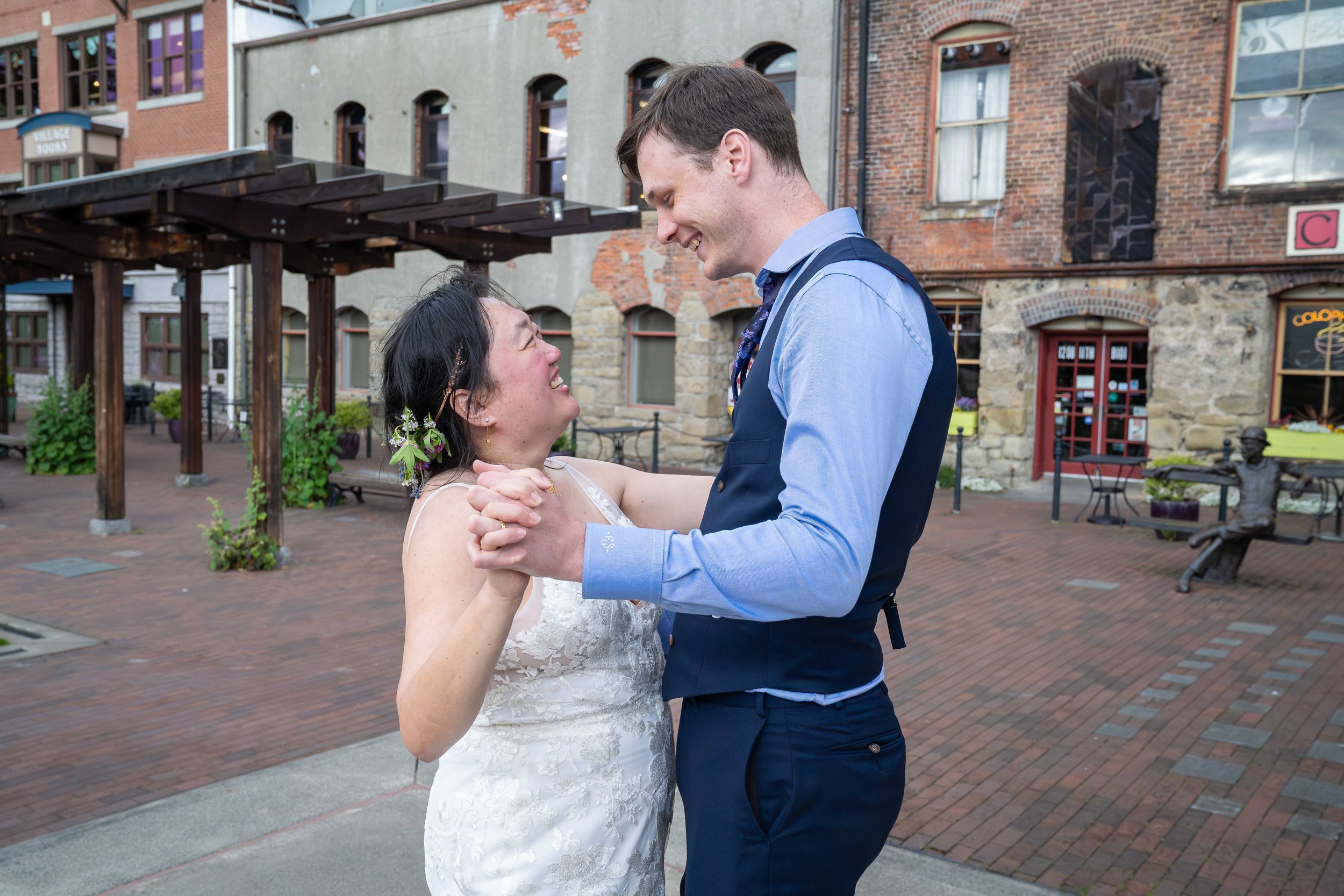 A bride and groom hold their first dance on the Village Green in historic downtown Fairhaven during their Bellingham elopement. They smile at one another and the brick-lined plaza and old stone buildings are visible behind them.