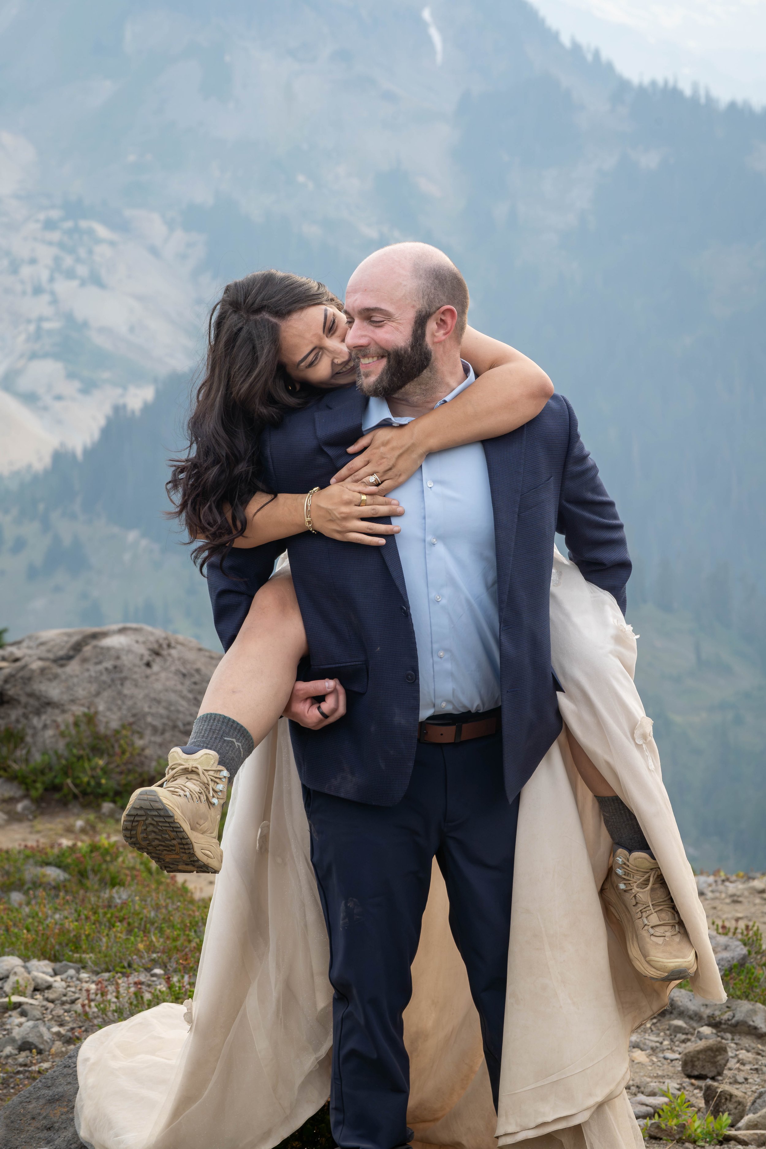 A bride jumps on her groom's back and puts her arms around his shoulders during a Mt. Baker elopement at Ptarmigan Ridge. They're standing in a rocky meadow with Table Mountain visible directly behind them.