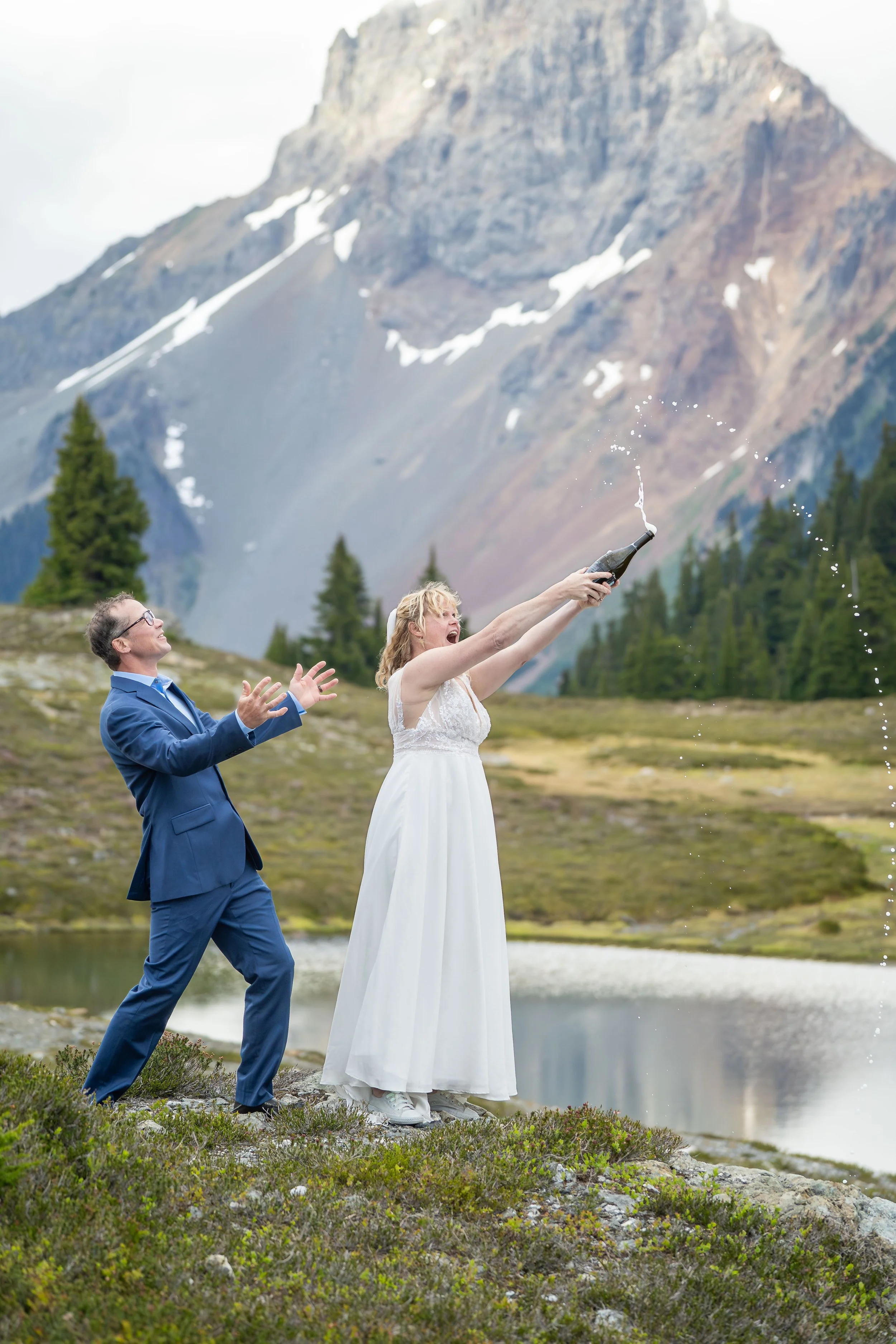 A bride in a wedding dress spraying a just-opened bottle of champagne as her groom looks on during their North Cascades elopement at Yellow Aster Butte. They are standing in front of an alpine lake with American Border Peak in the background.