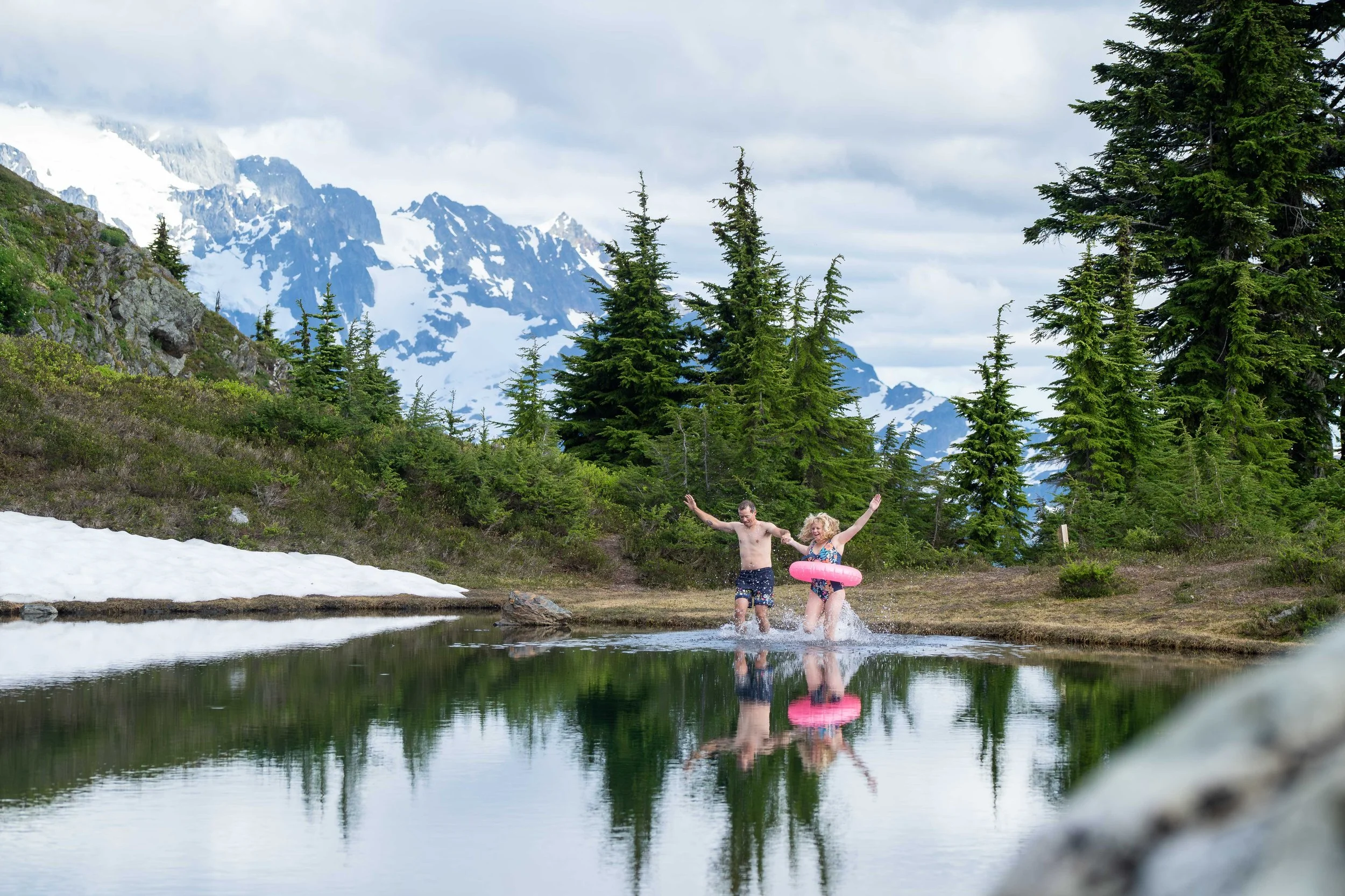 A bride and groom running into an alpine lake in the North Cascades with Mount Shuksan visible behind them. There's snow on the edge of the lake and it's surrounded by subalpine meadow.