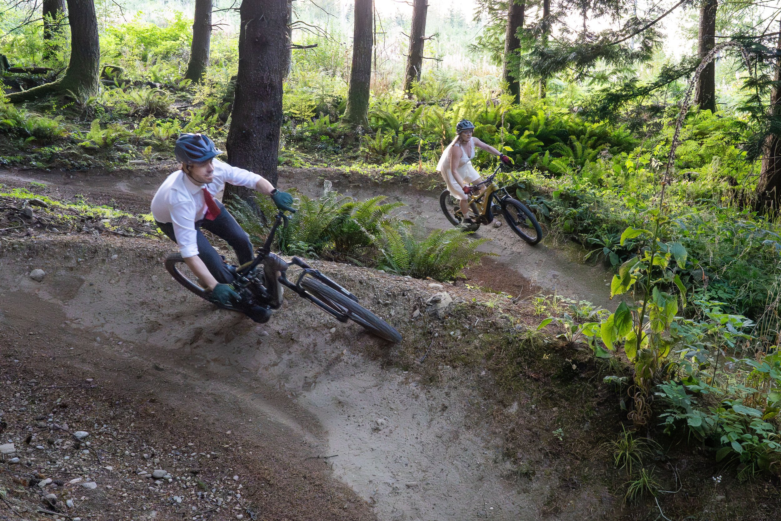 A bride and groom riding mountain bikes side-by-side on a large berm in a dual slalom course on Galbraith Mountain in Bellingham. The bride is wearing a wedding dress and the groom is wearing a suit.