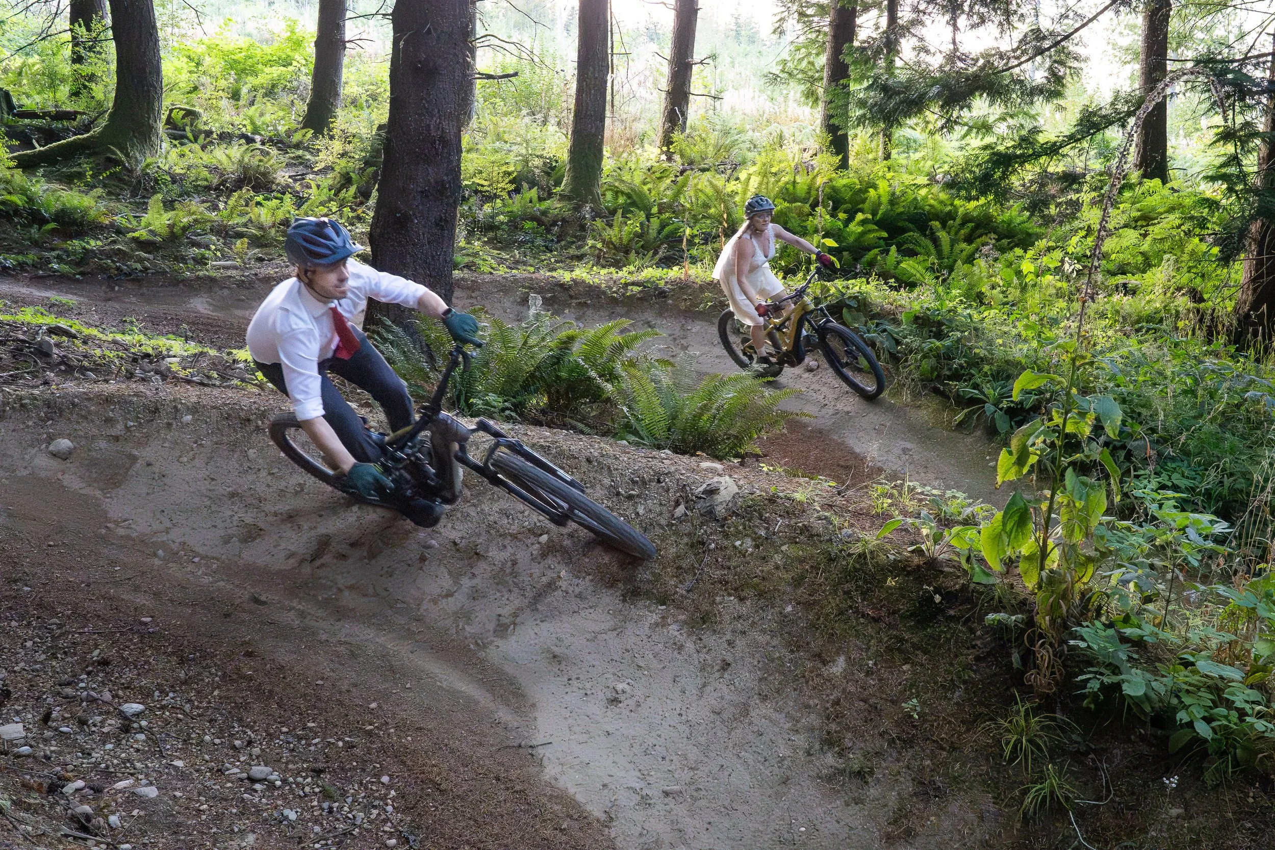A bride and groom riding mountain bikes down Stinky Dual-uxe, a dual slalom course, while wearing full wedding clothes during a mountain biking elopement photoshoot on Galbraith Mountain, in Bellingham, Washington.