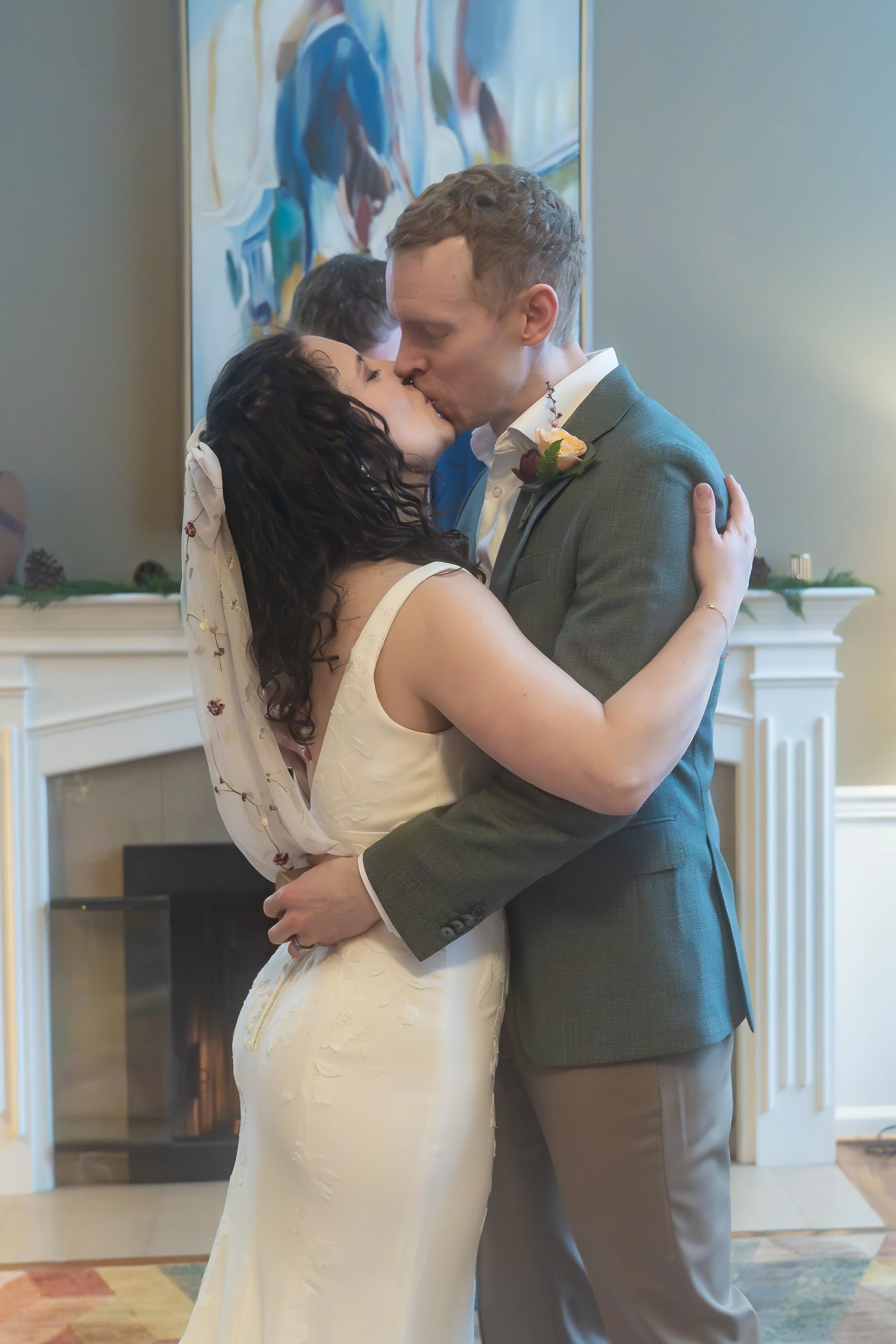 Photo of a couple kissing at the end of their wedding ceremony. The woman is wearing a white wedding dress and the man a gray suit, and they're in a living room with a fireplace behind them.