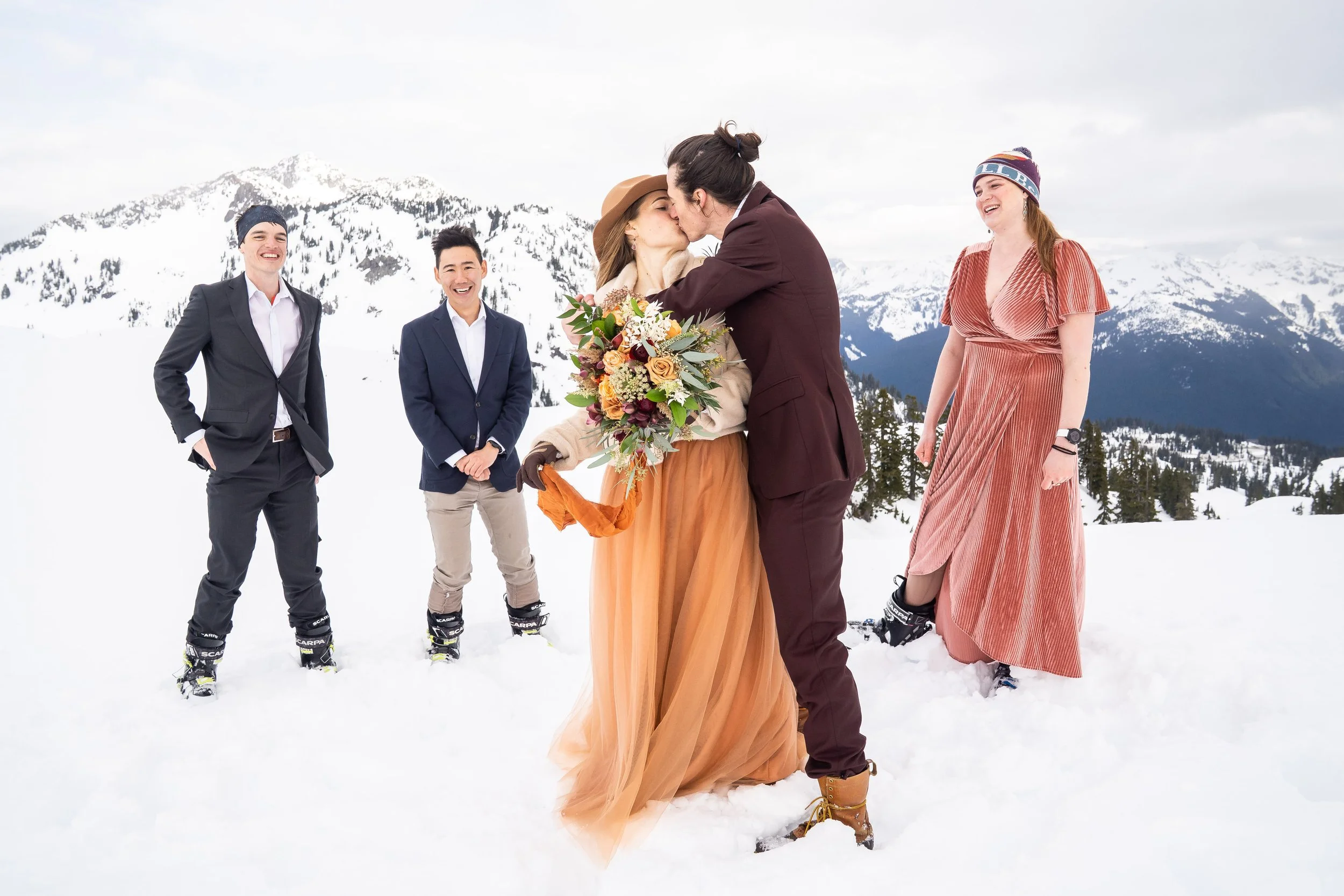 A photo of a small wedding party standing in the snow in ski boots. The bride and groom are in the center kissing and the bridesmaid and groomsmen are arranged around them.