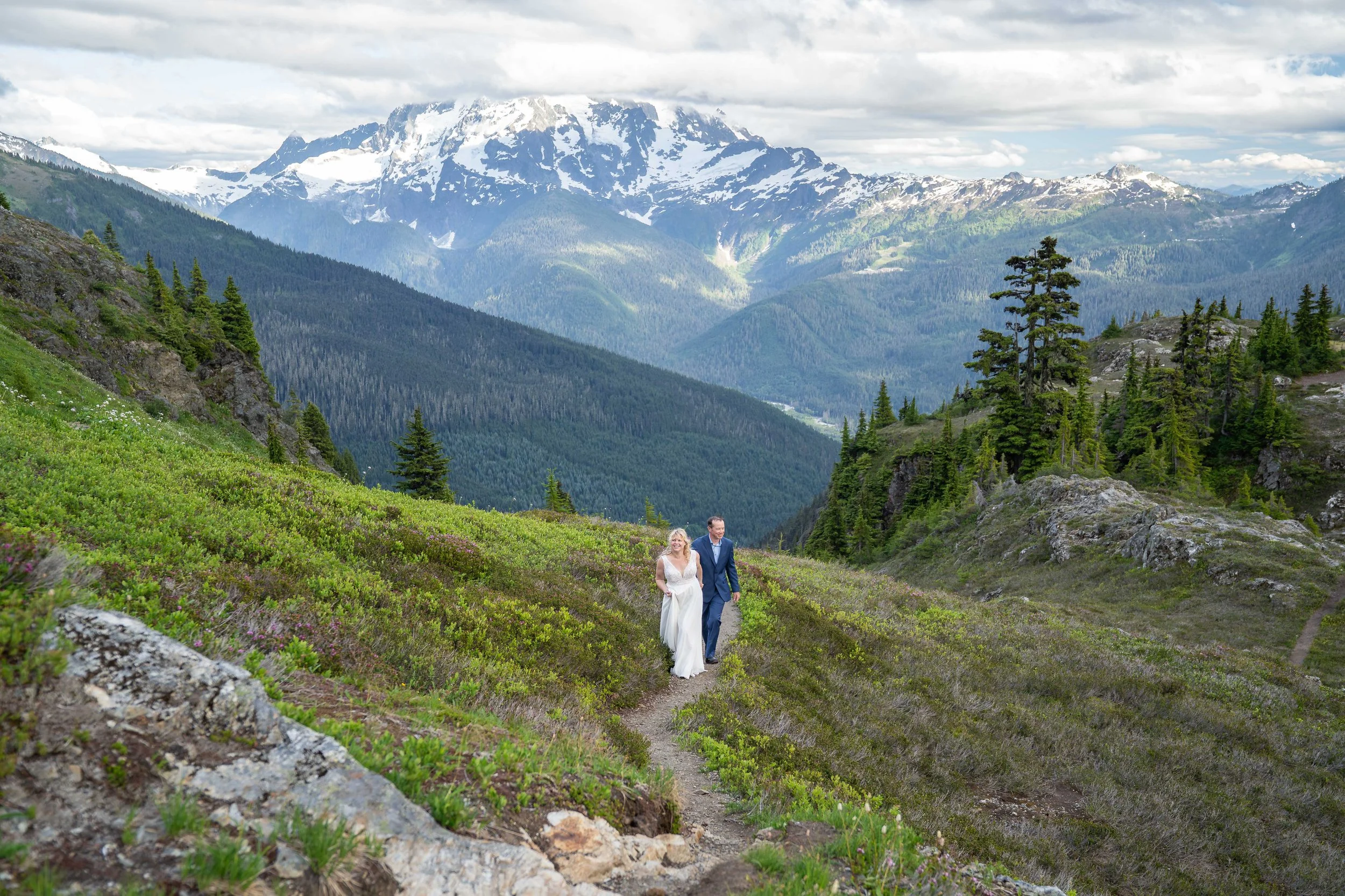 A bride and groom holding hands as they walk through a subalpine meadow near Yellow Aster Butte and High Divide, in the North Cascades, with Mt. Shuksan visible in the background.