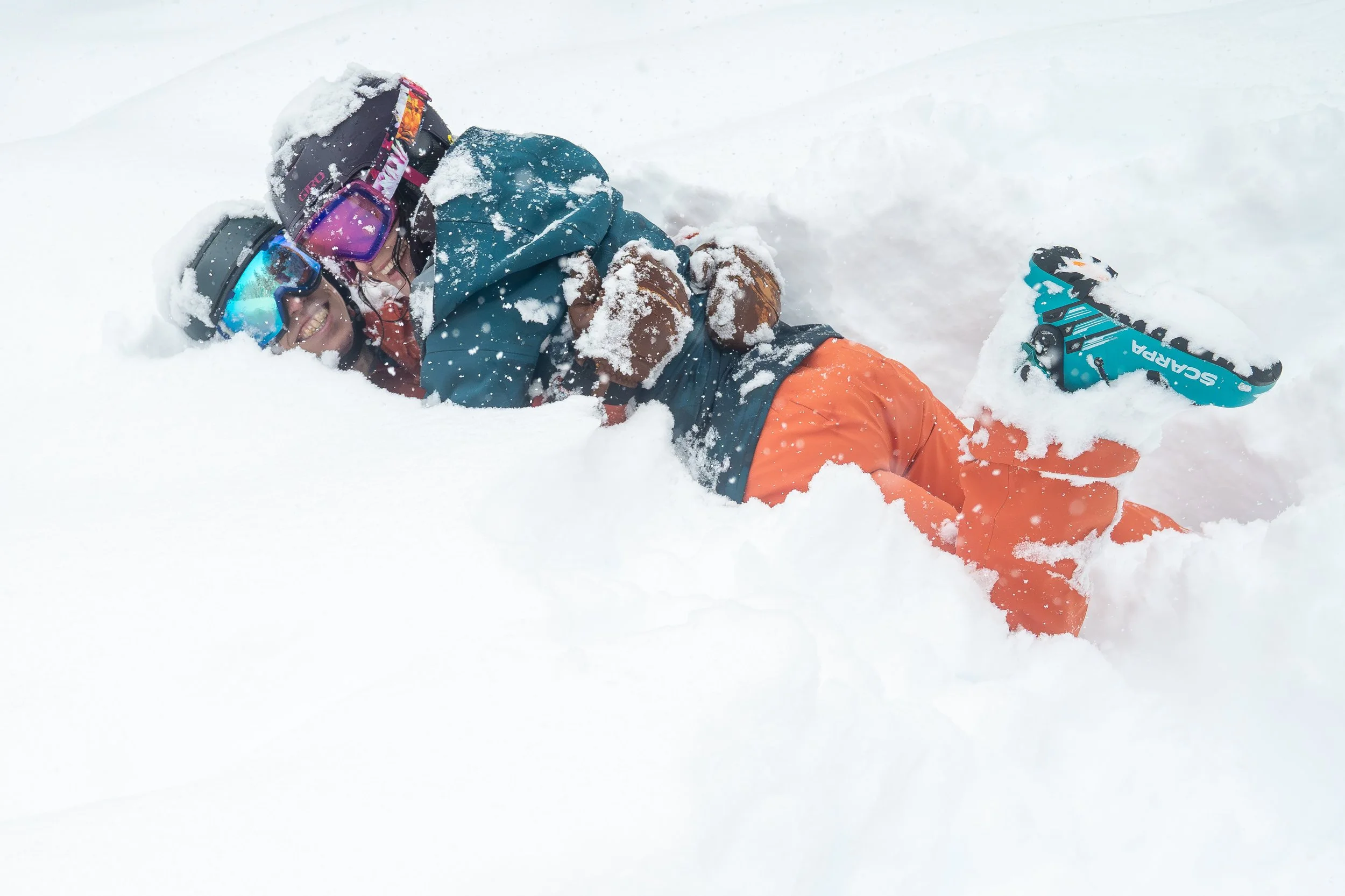 Photo of a man and woman in ski clothes lying in deep snow while smiling and laughing. The woman is on top of the man with her ski boot kicked up in the air.
