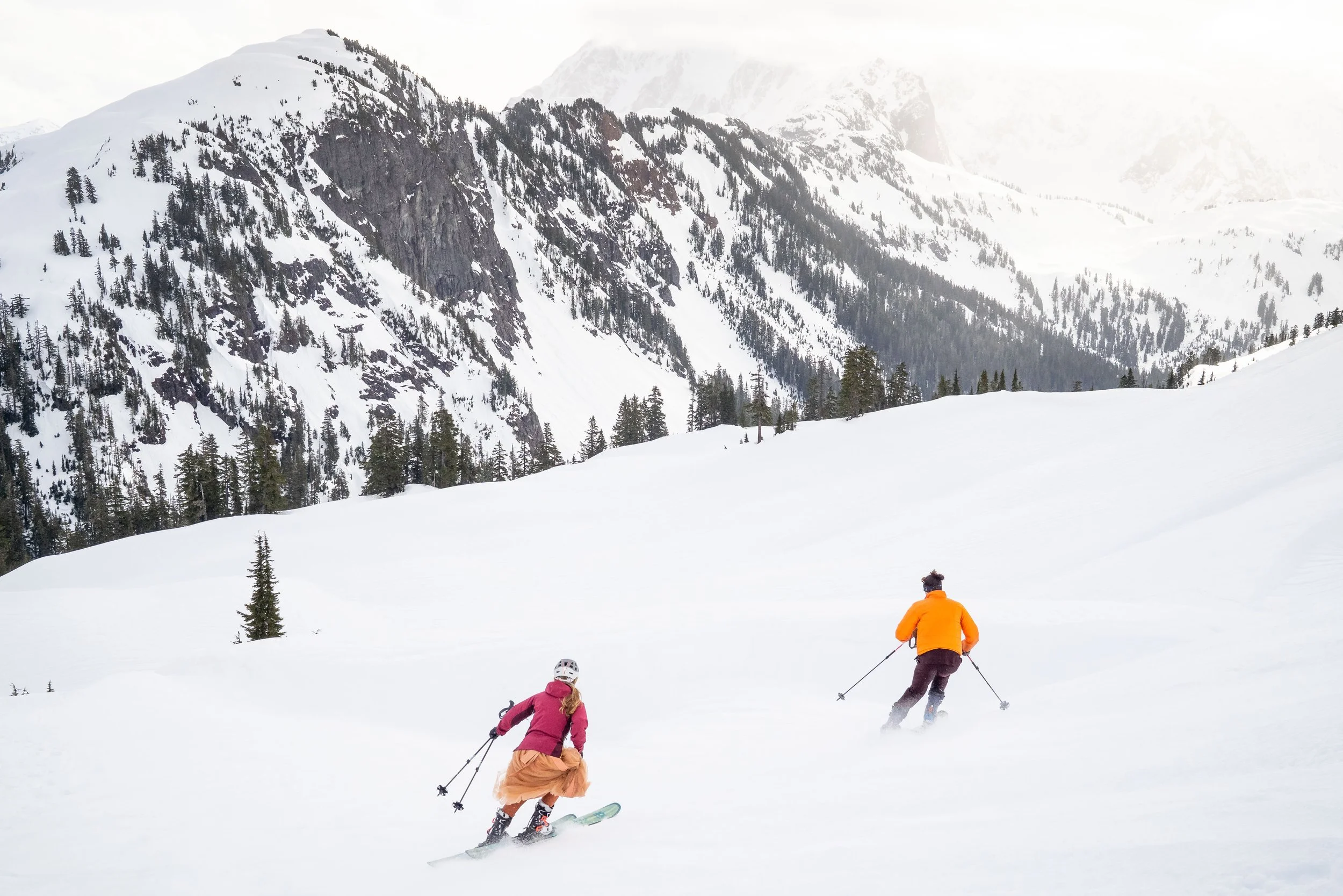 A photo from behind of a bride and groom skiing in powder with Mt. Shuksan in front of them. The woman is wearing an orange wedding dress and the man is wearing an orange jacket over a suit.