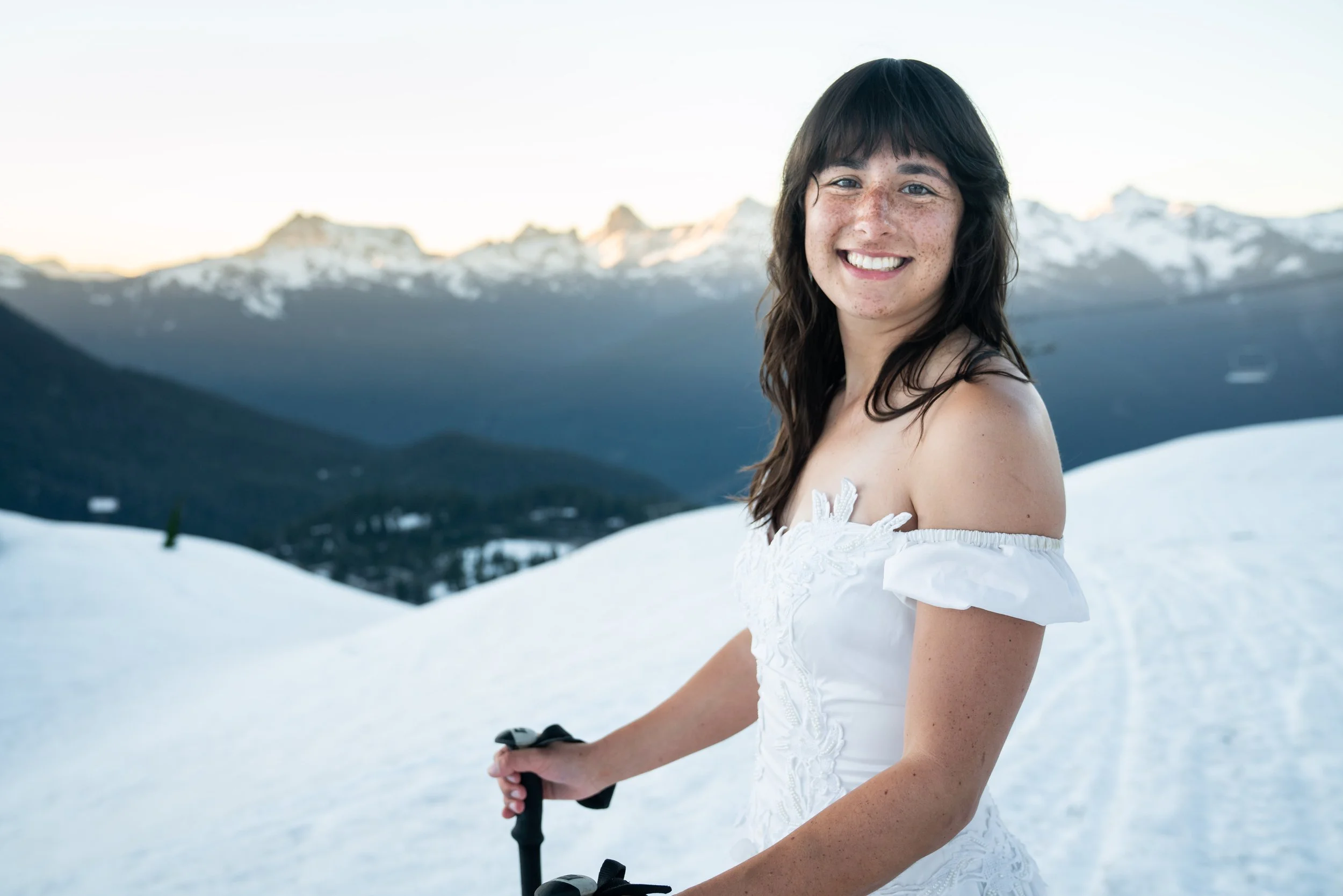 A photo of a woman in a vintage wedding dress standing in the snow and smiling at the camera. She's holding ski poles and there are mountains at sunset in the background.