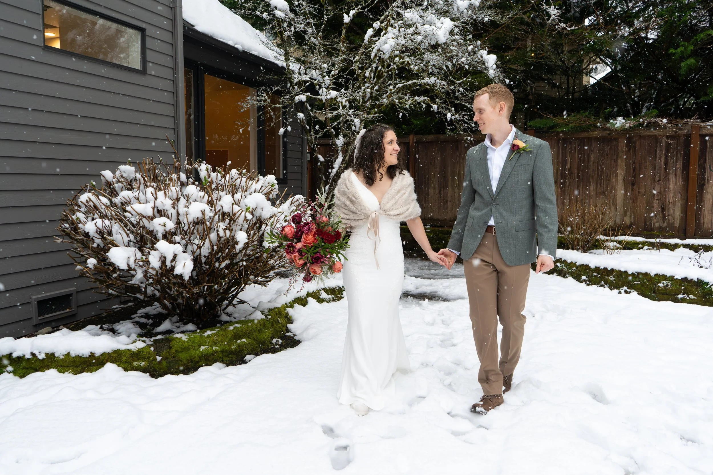 Photo of a woman in a white wedding dress and a man in a gray suit holding hands and looking lovingly at each other while they walk towards the camera. They're in a snowy backyard garden surrounded by trees.