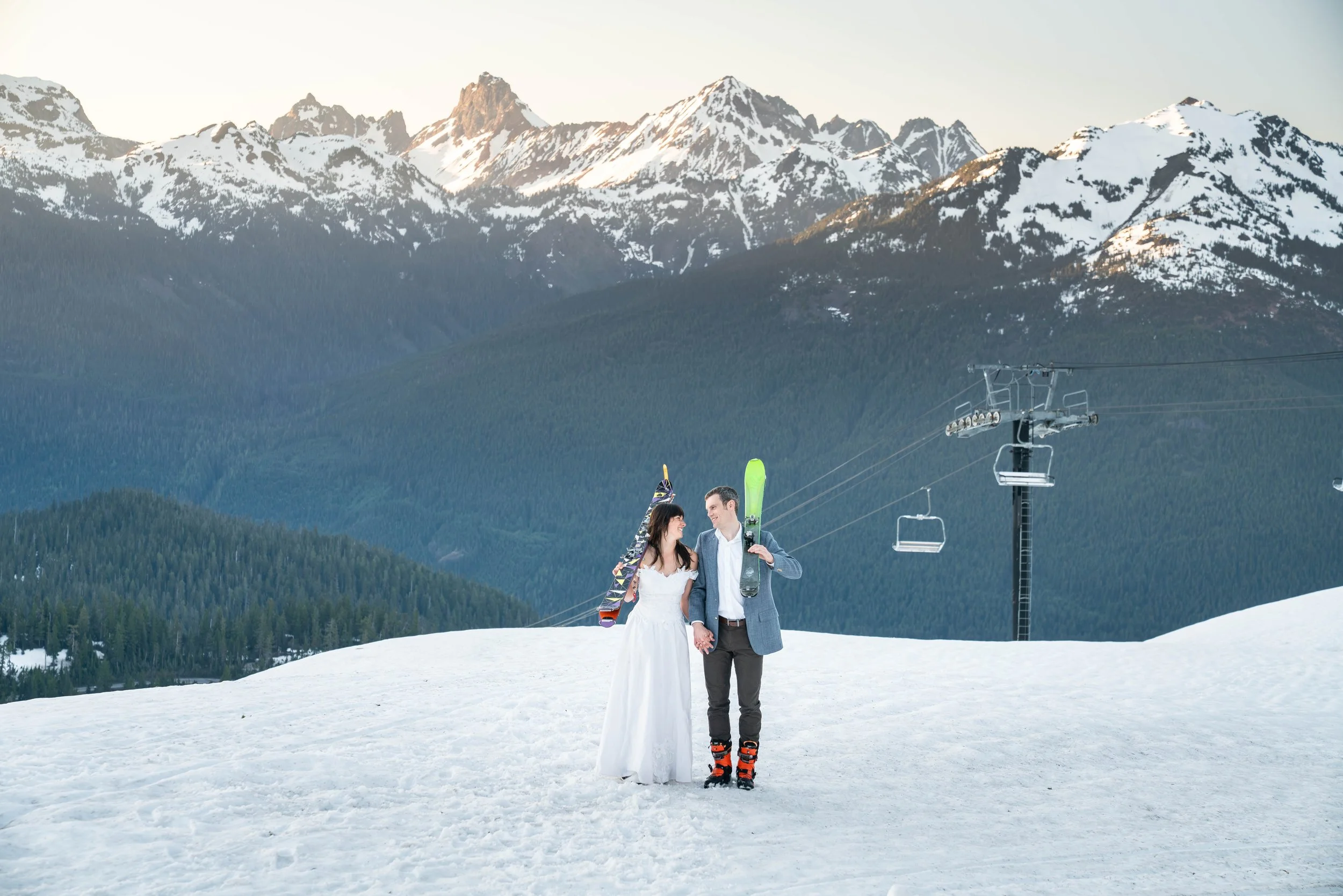 A photo of a couple in vintage wedding clothes and ski boots standing in the snow and holding skis over their shoulders. Snowy mountains at sunset are visible in the background.