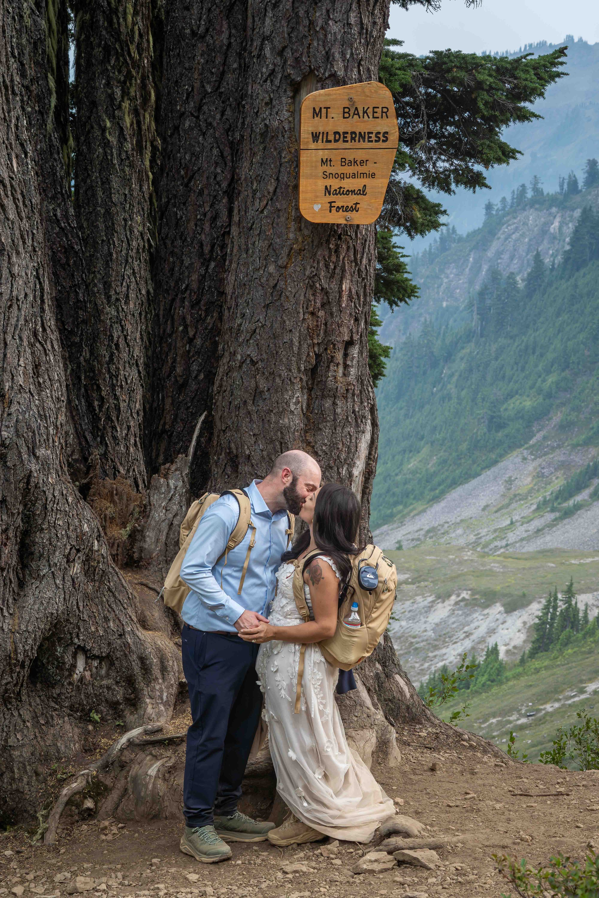 A bride and groom kiss beneath the Mt. Baker Wilderness sign on the Ptarmigan Ridge trail during their Mt. Baker elopement. They are wearing wedding clothes and matching backpacks, an the sign is high above them on a large fir tree.