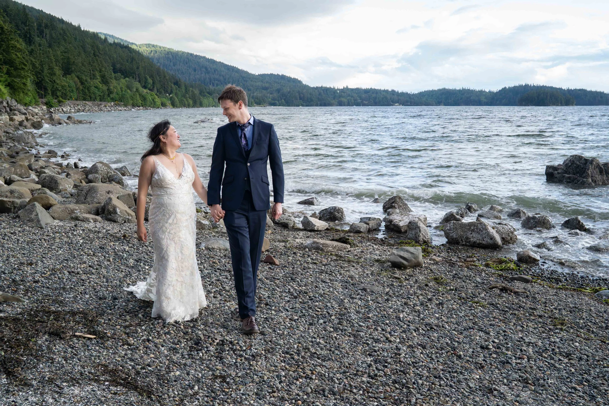 A bride and groom walking together on a pebbly beach at Teddy Bear Cove at sunset after their Bellingham, Washington elopement. The water is visible behind them as they walk towards the camera.