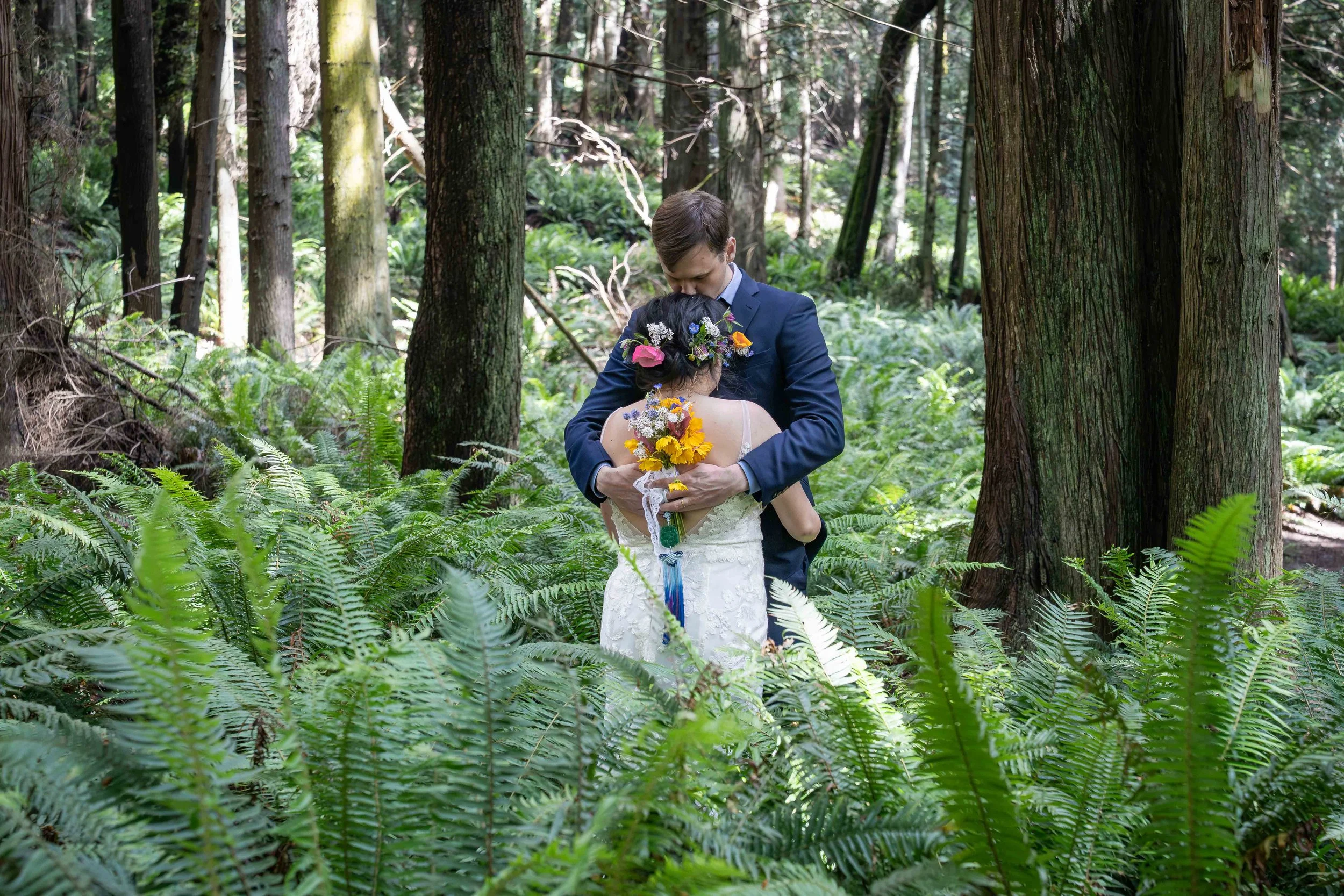 A bride and groom in wedding clothes embrace while surrounded by lush ferns and large trees on the Fragrance Lake Trail in Larrabee State Park, just before their wedding ceremony.
