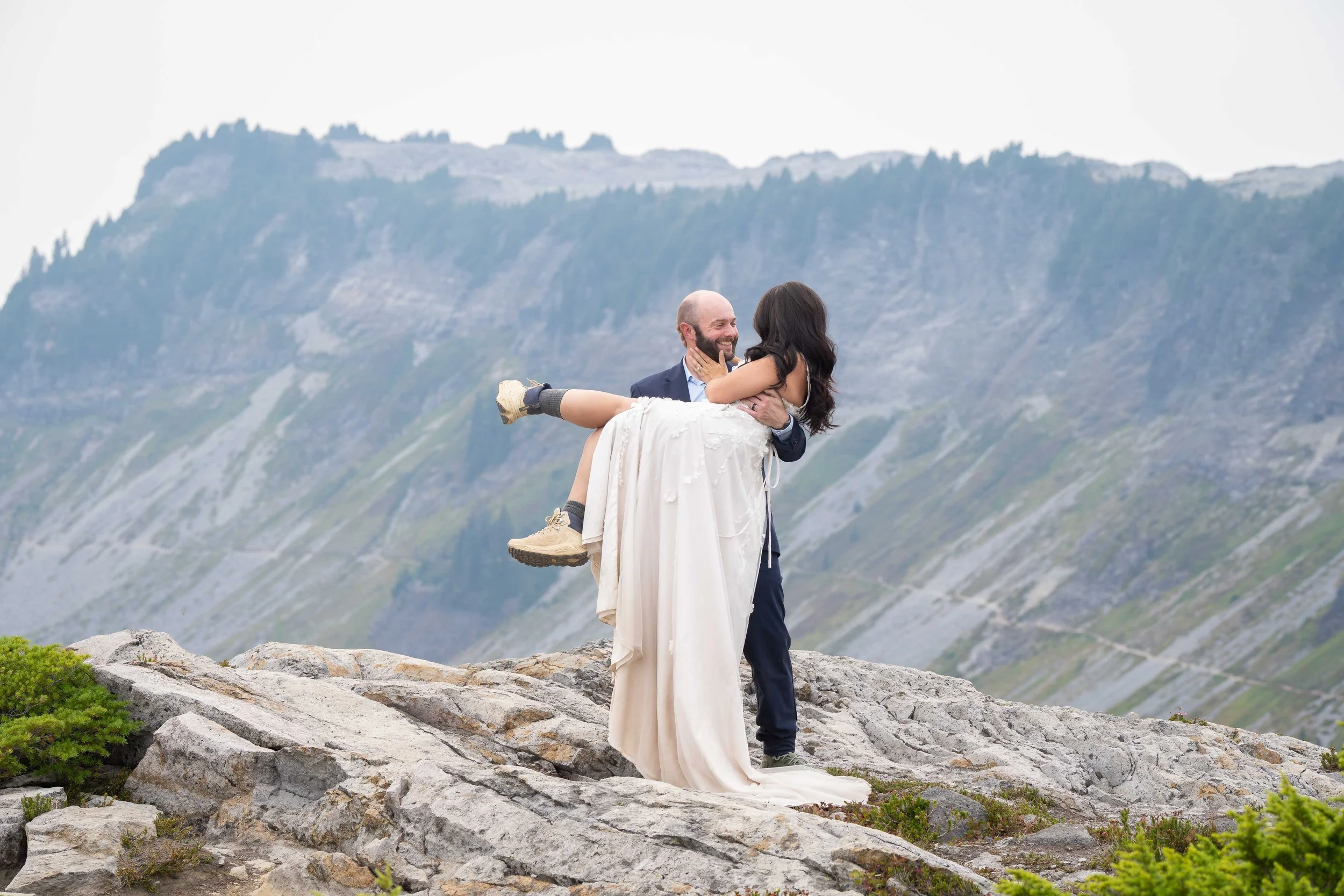 A groom holding his bride during an Artist Point elopement. The bride is wearing a wedding dress and they're standing on a rocky overlook with Table Mountain in the background.