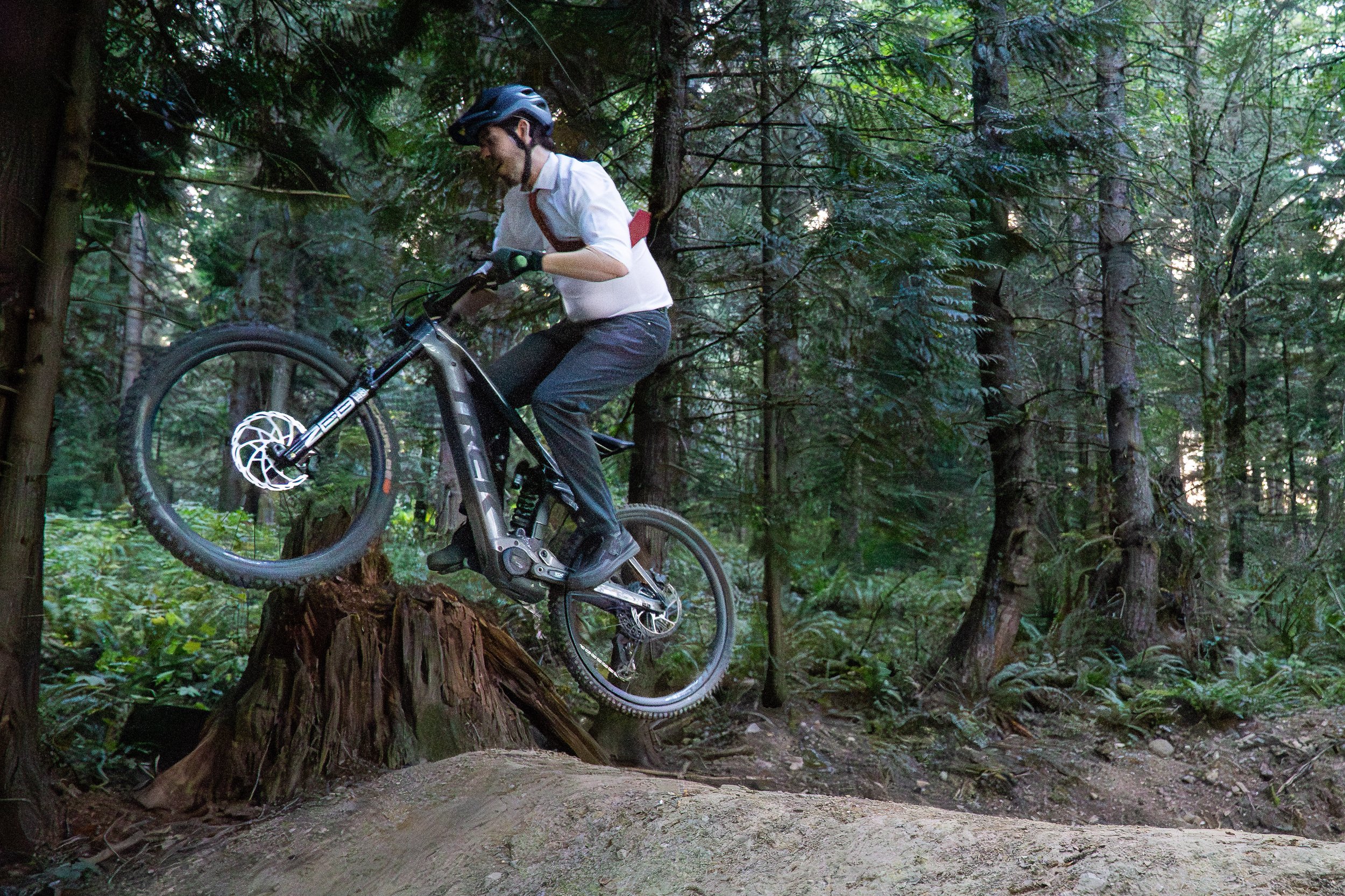A groom catching air with his e-mountain bike off a jump during a mountain biking elopement photoshoot on Galbraith Mountain, in Bellingham, Washington. he is wearing a wedding suit and tie.