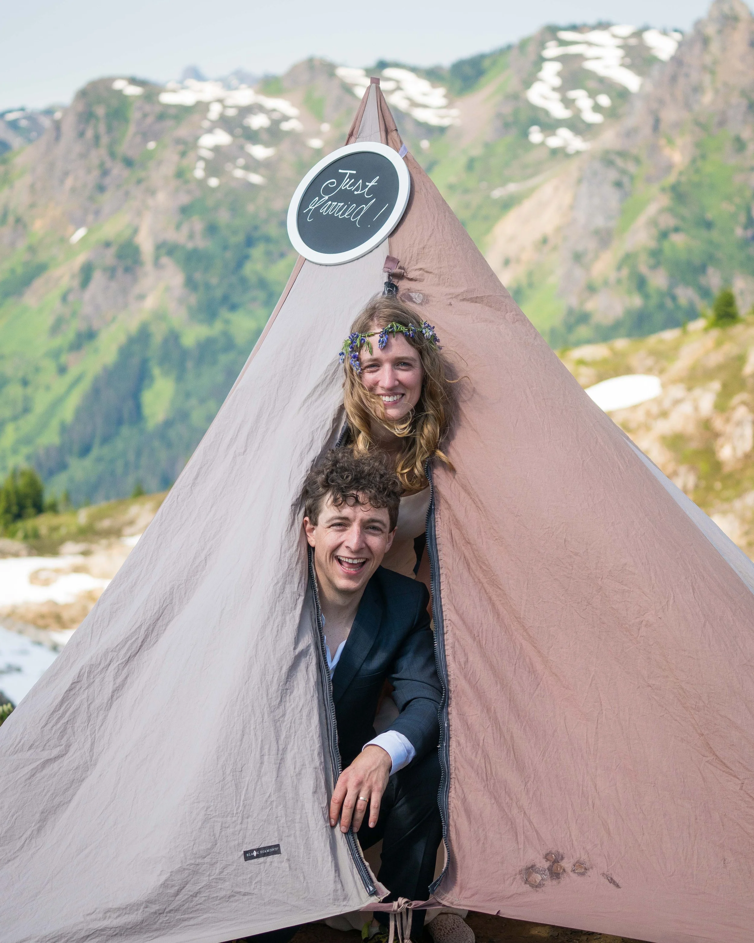 A bride and groom poke their heads out of their tent after being exchanging vows during a North Cascades elopement at Yellow Aster Butte. There's a chalkboard sign hanging on the tent that says "Just Married!"