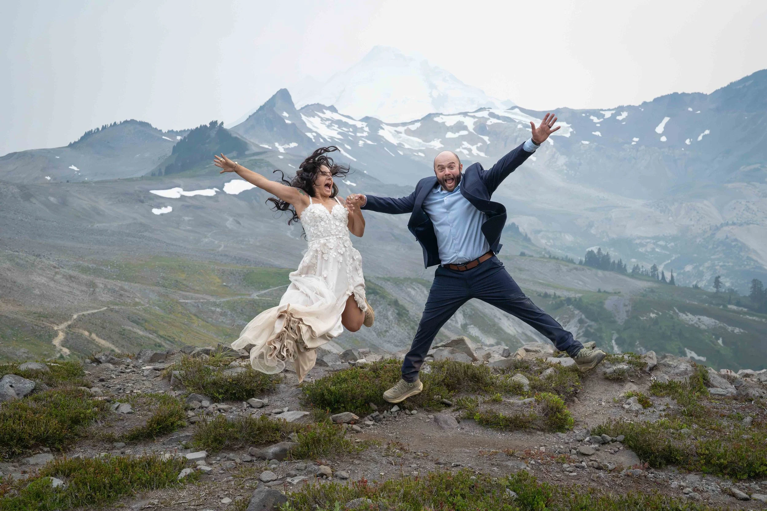 A bride and groom in wedding clothes jumping in the air together during their Ptarmigan Ridge elopement. The Ptarmigan Ridge trail, Coleman Pinnacle, and Mt. Baker are visible behind them at sunset.