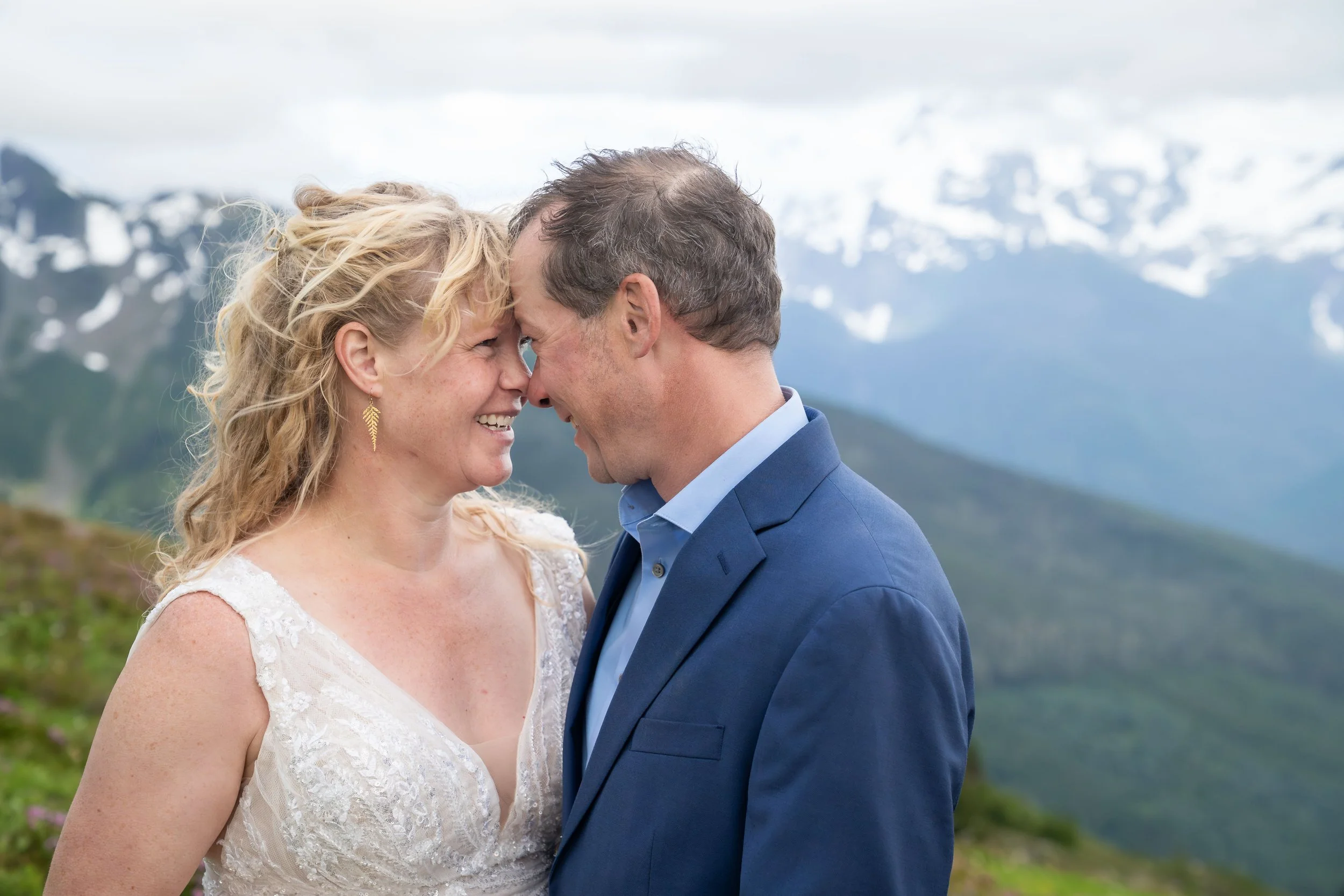 A bride and groom in wedding clothes touching their foreheads and noses together during a North Cascades sunset elopement at Yellow Aster Butte. They are standing in a subalpine meadow and Mount Baker is visible in the background.